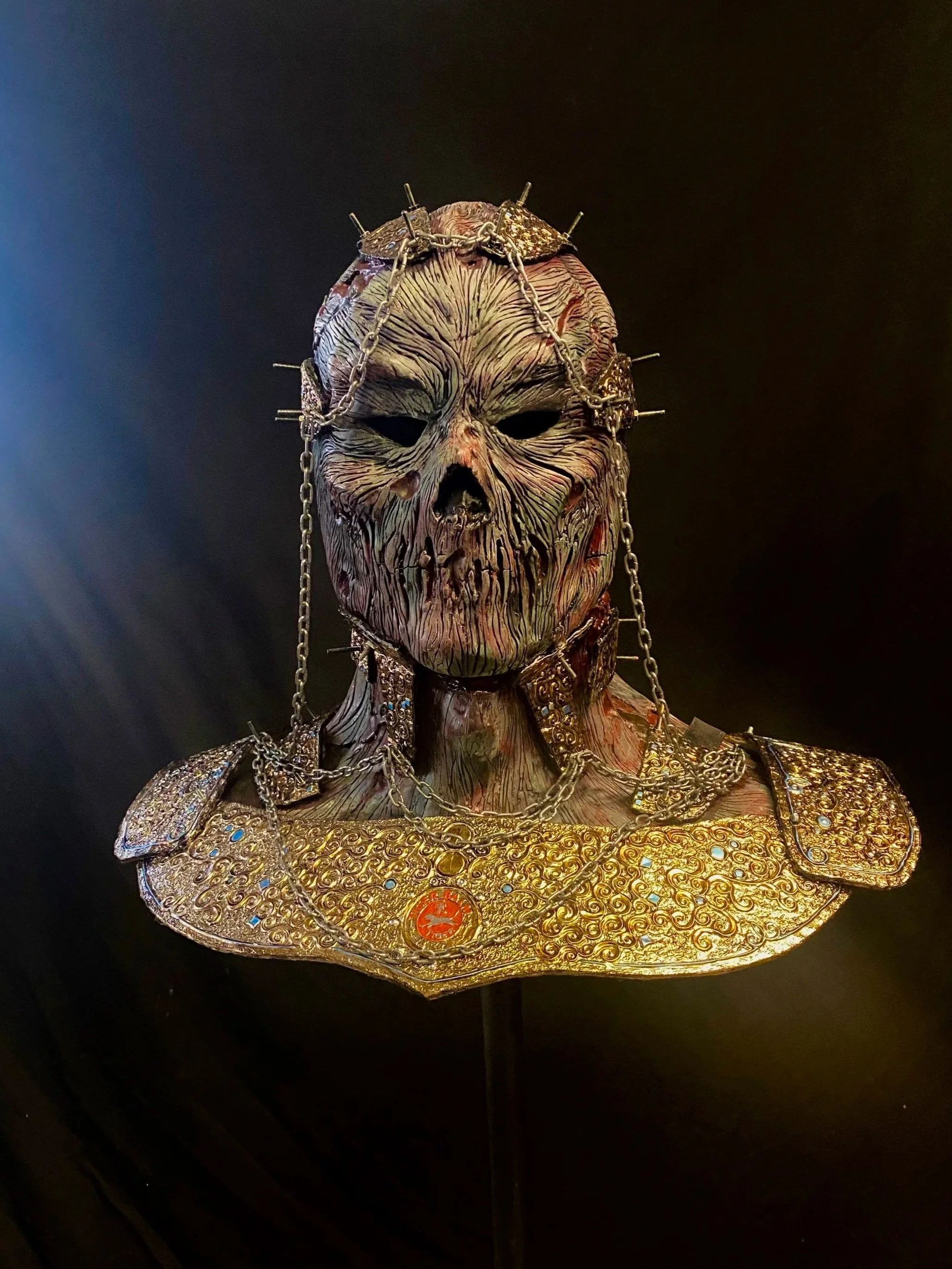 A mask resembling a scary, wrinkled face with dark eye holes, adorned with chains and a golden shoulder piece decorated with intricate patterns and embedded gemstones.