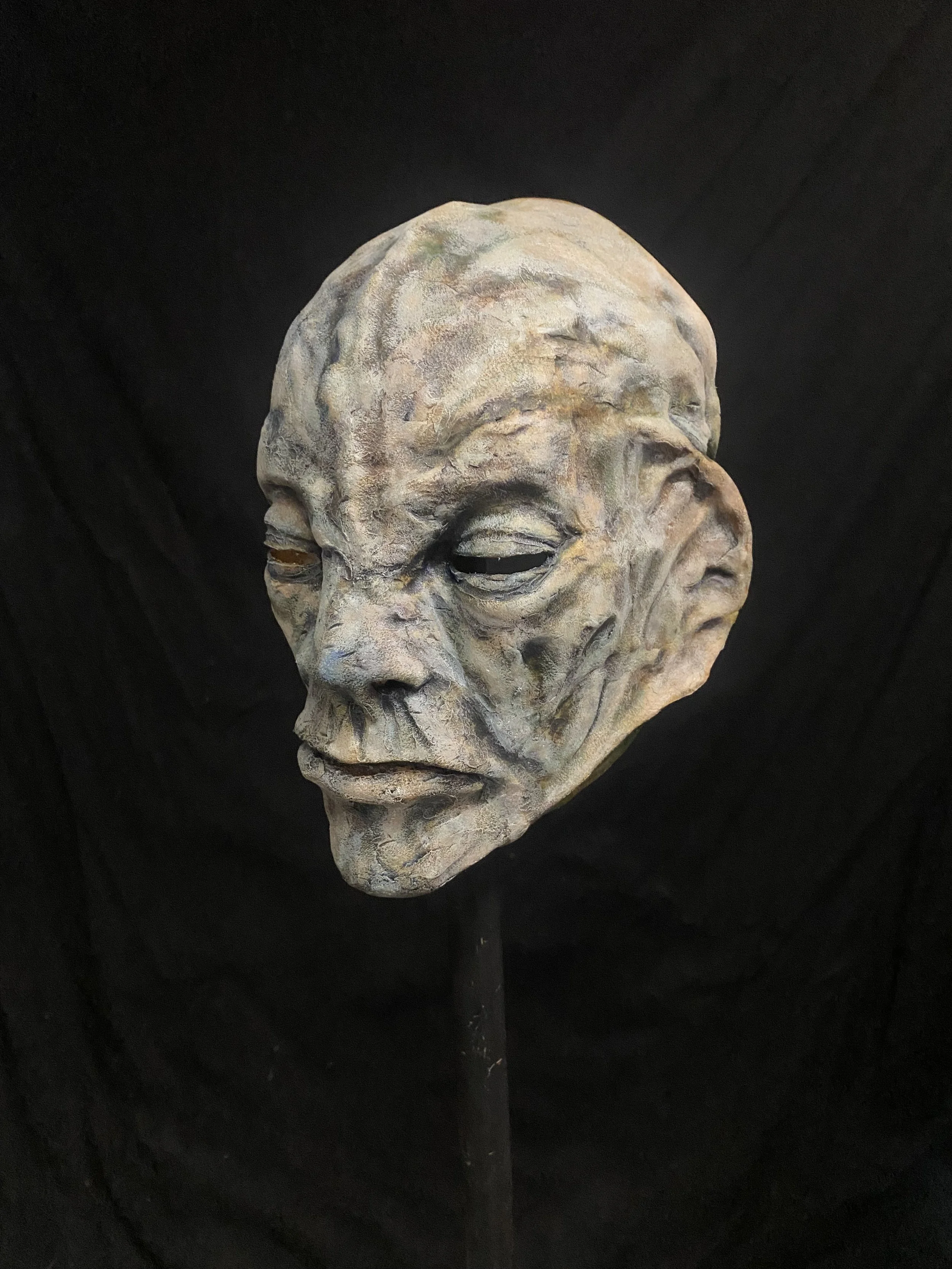 A sculpture of a weathered, aged human face on a stand, with closed eyes and a contemplative expression, against a black background.