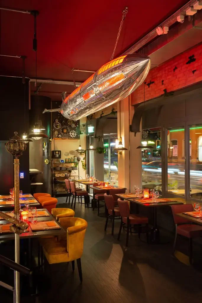 Interior of a restaurant with a rocket-shaped light fixture is suspended from the ceiling, warm lighting, tables set with glassware and candles, and large windows showing city lights.