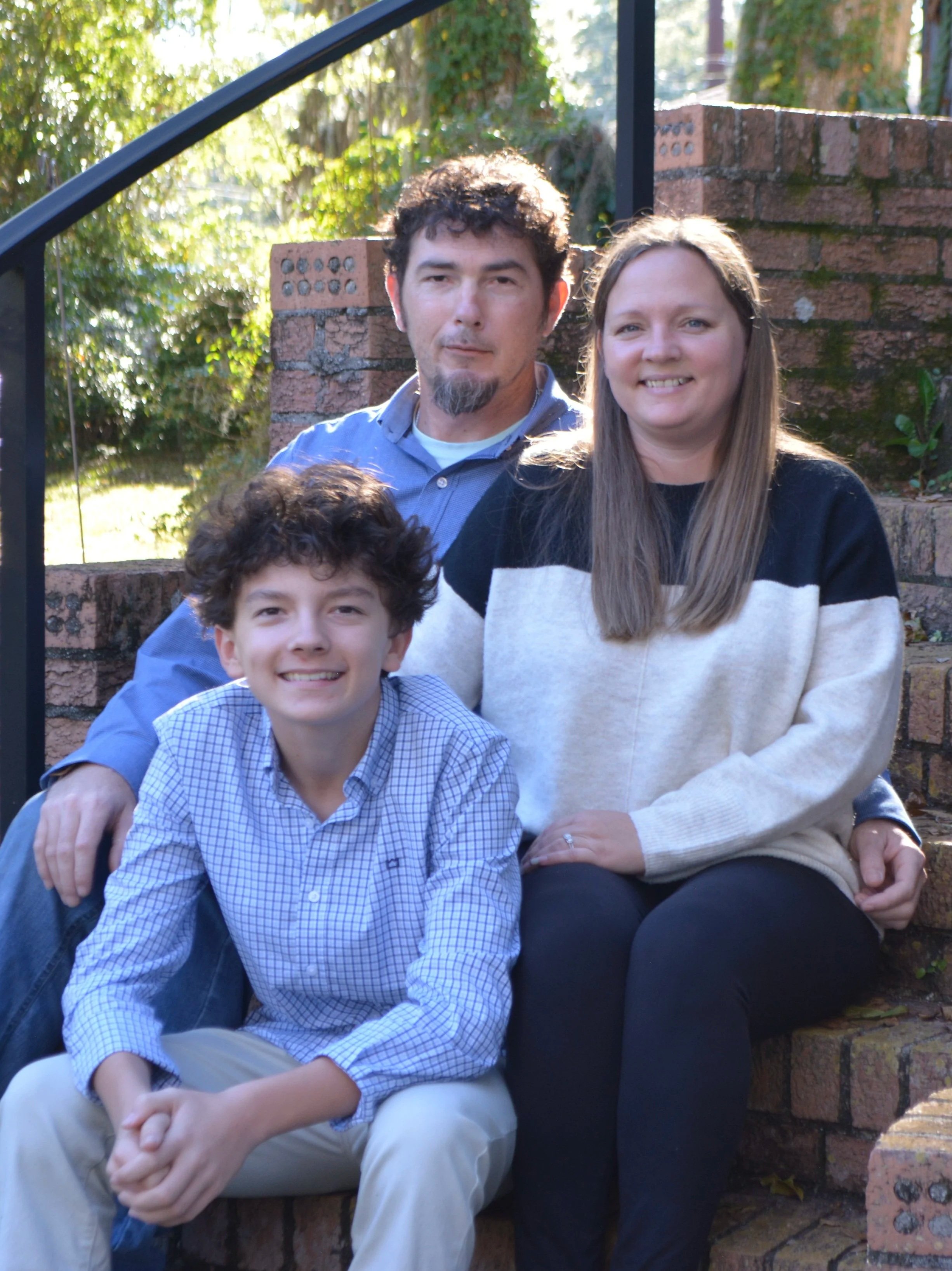 Three people sitting on brick stairs outside, with green trees and sunlight in the background. They are smiling and posing for the photo.