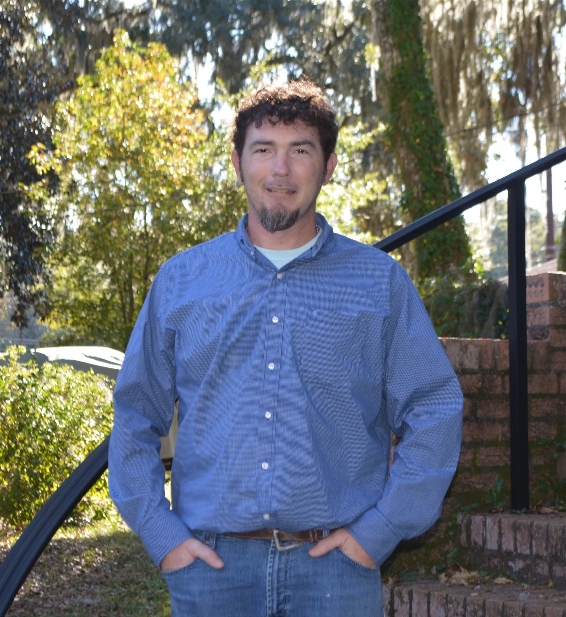 A man with dark curly hair and a facial beard, wearing a blue button-up shirt, standing outdoors with trees and sunlight in the background.
