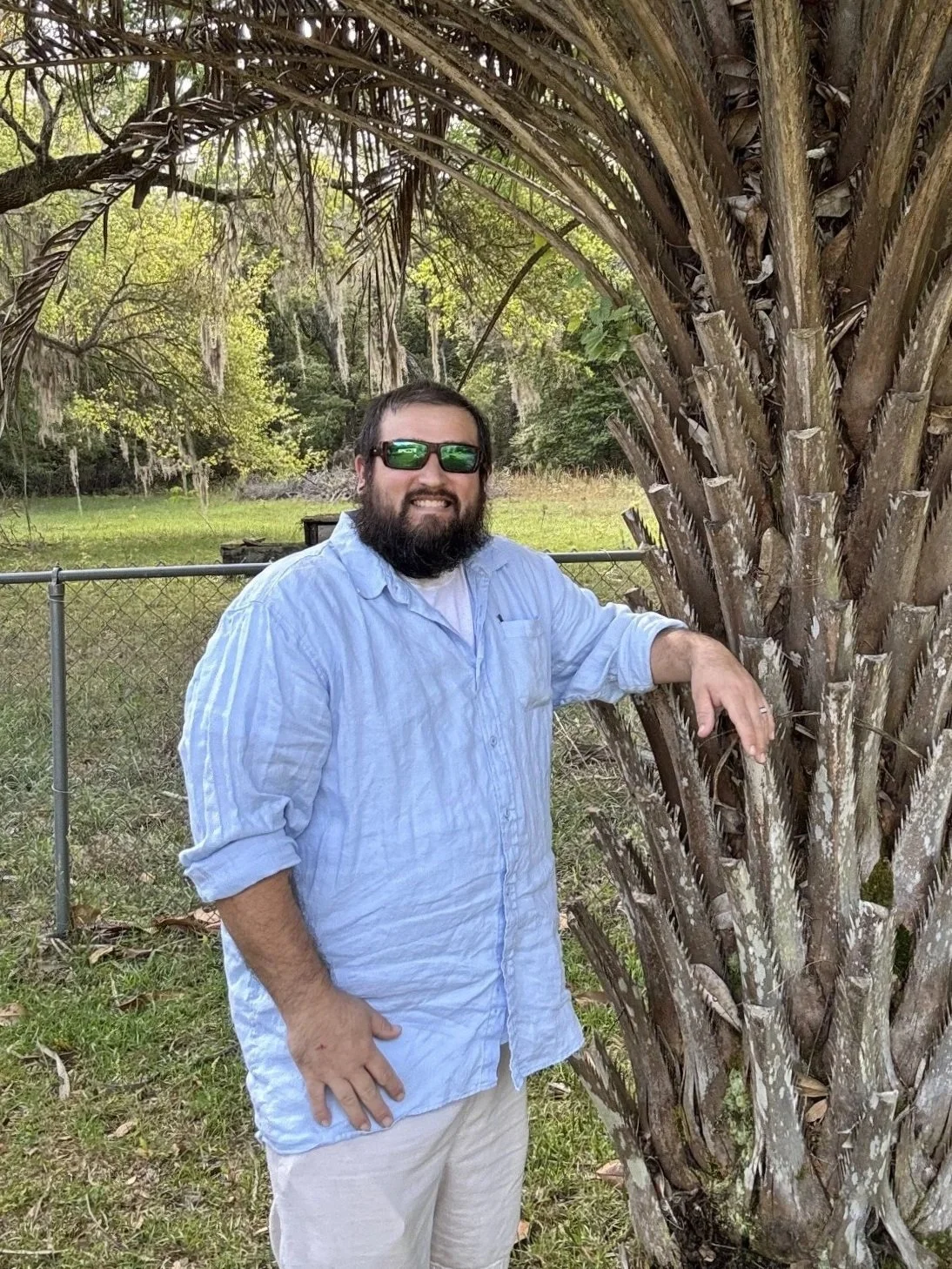 A man with a beard wearing sunglasses, a light blue shirt, and beige pants, standing outdoors next to a large palm tree, smiling, with a grassy yard and trees in the background.