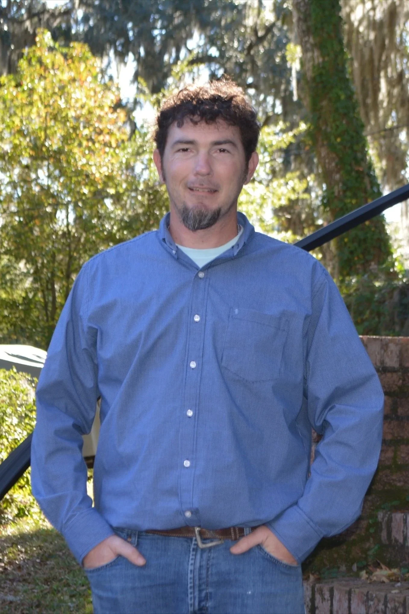 A man with dark, curly hair and a goatee, wearing a blue button-up shirt, standing outdoors with trees and foliage in the background.