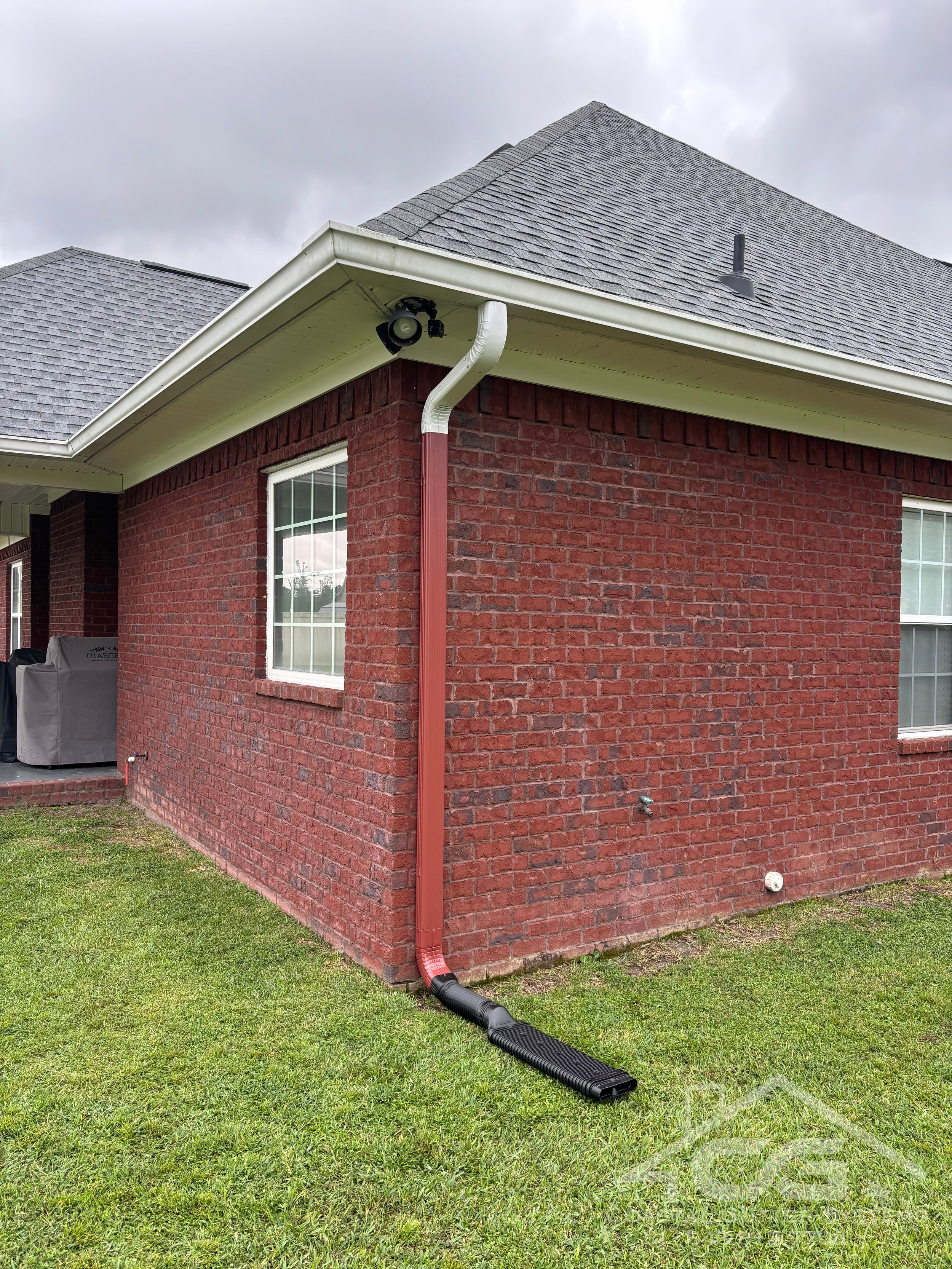 A brick house with a grey shingle roof and white trim, featuring a window and a downspout connected to a black underground drainage system, with a grassy yard in the foreground and a cloudy sky above.