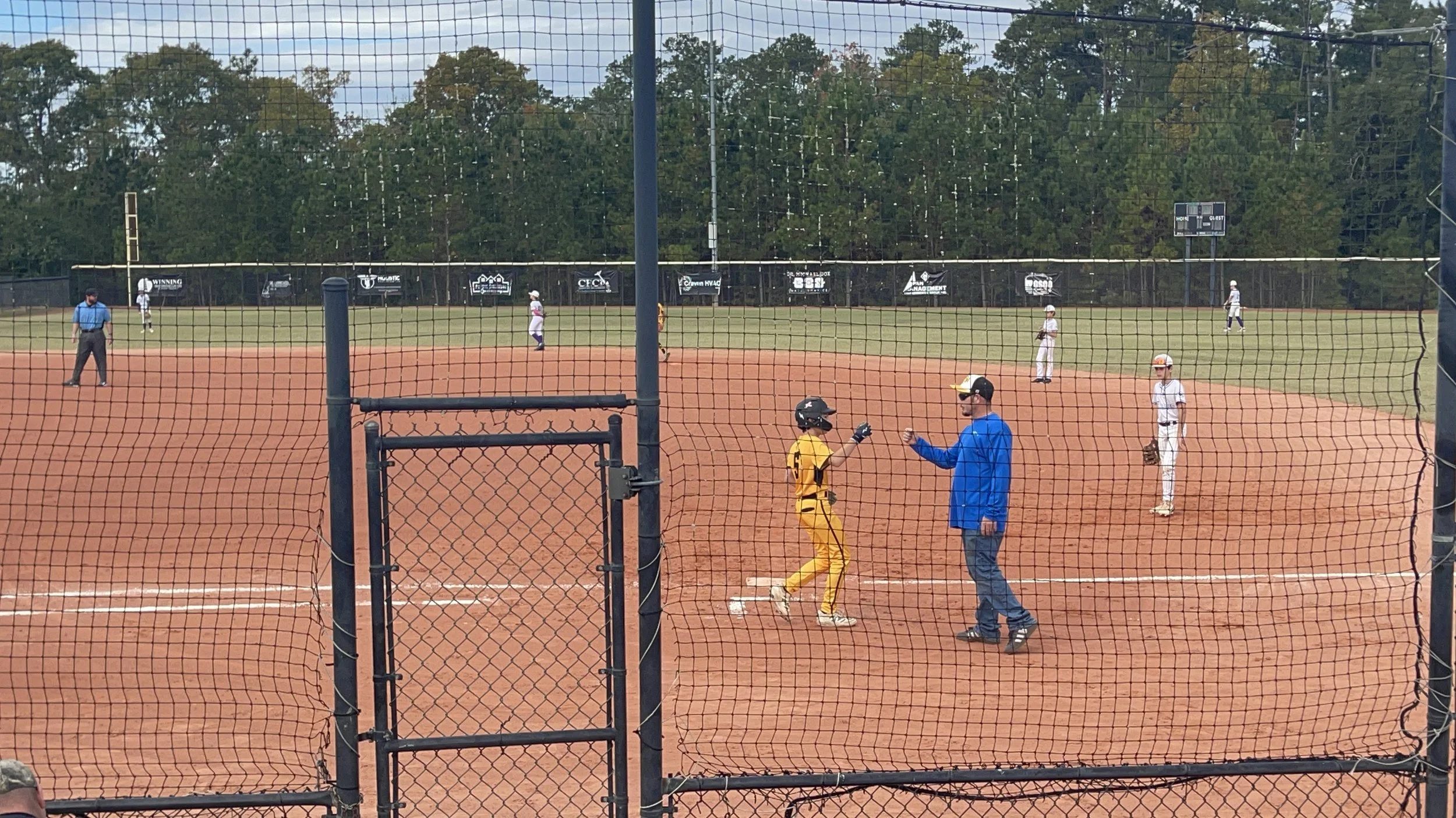 A baseball game with players on the field, an umpire, and coaches near home plate on a dirt infield, viewed through a protective fence.