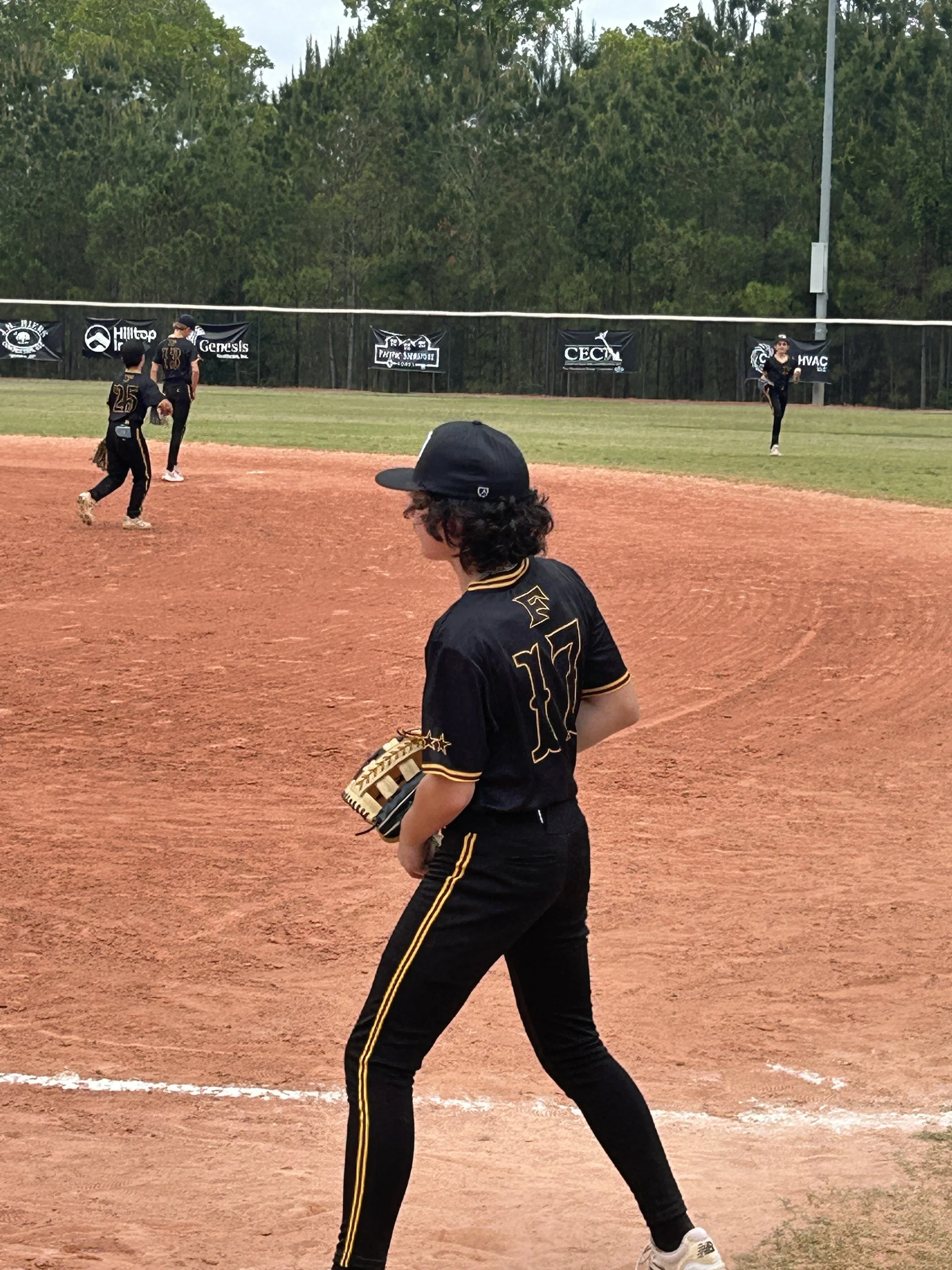 A group of young baseball players in black uniforms with yellow accents on a baseball field during daytime. One player in the foreground with curly hair and a cap is holding a baseball glove. Other players are in the background on the field, with some preparing to catch or throw the ball. The field has a dirt infield and grass outfield, with trees and advertising banners along the outfield fence.