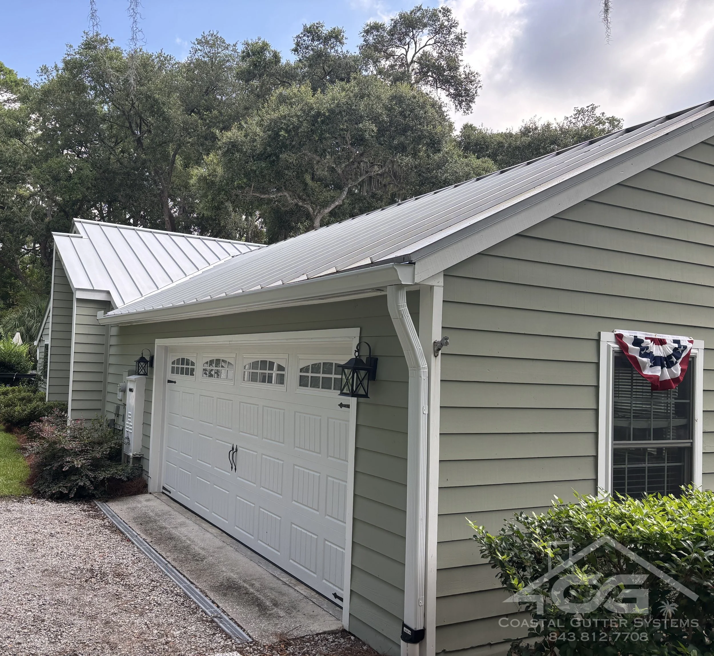 A green house with siding and a metal roof. House features a new white seamless gutter with a downspout over the garage. Landscaping and trees in the background.