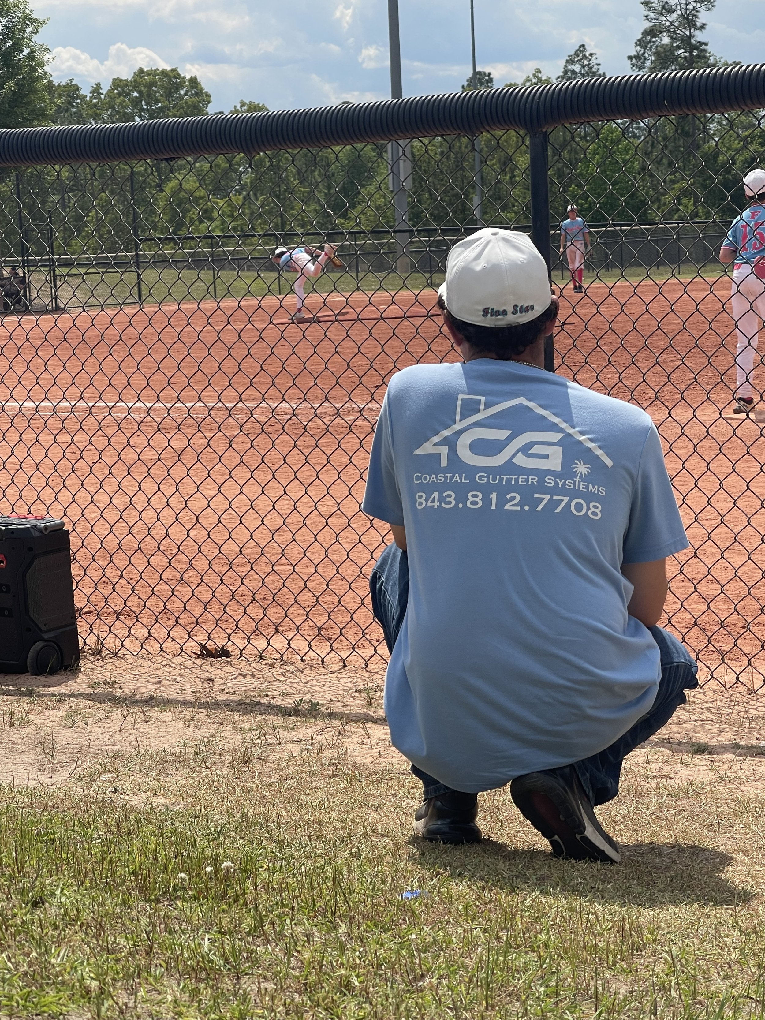 A person in a blue shirt and white cap watching a baseball game from outside the fence, with players on the field and a partly cloudy sky.