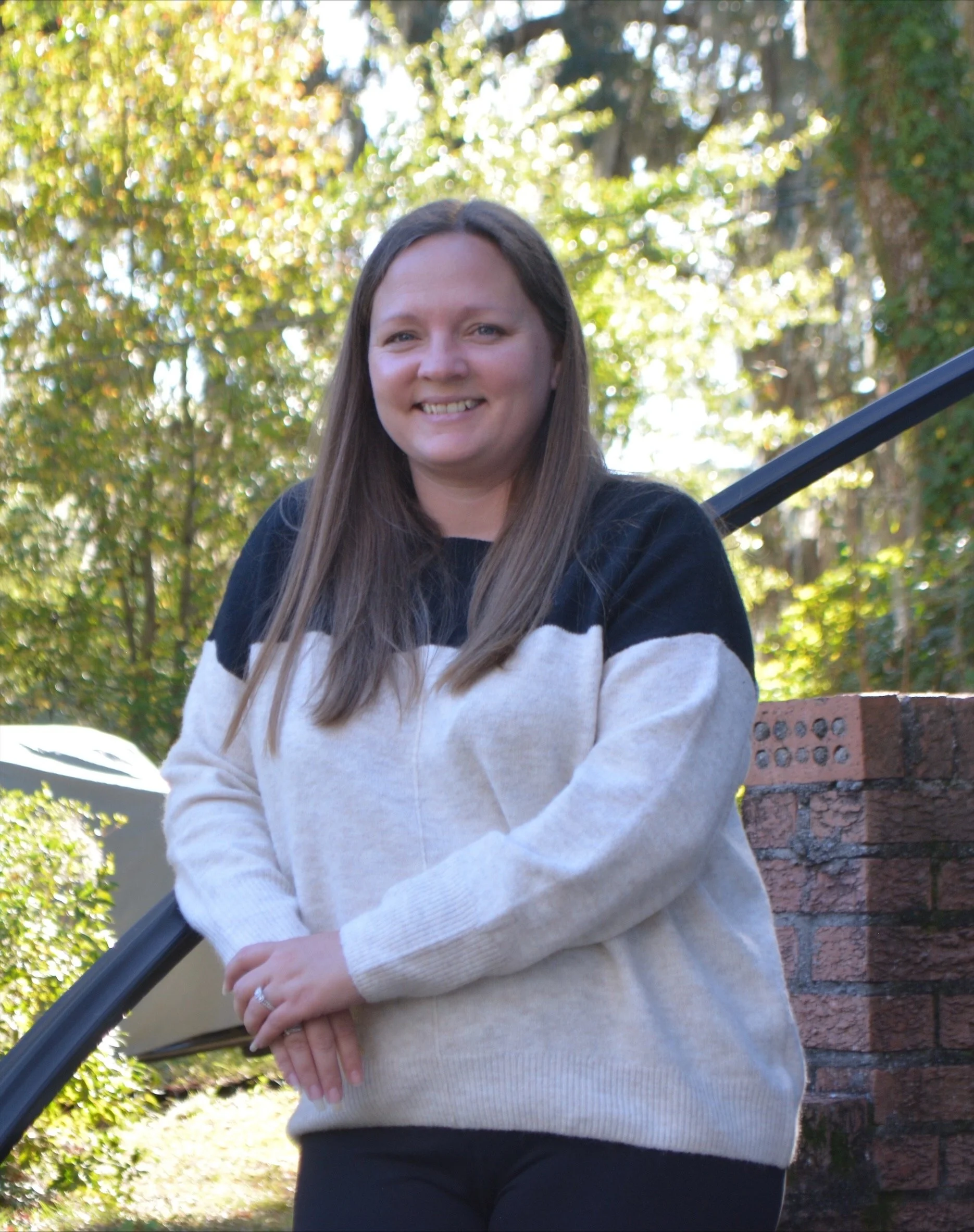 A woman with long brown hair smiling outdoors on a sunny day, standing in front of trees, a brick wall, and a metal railing.