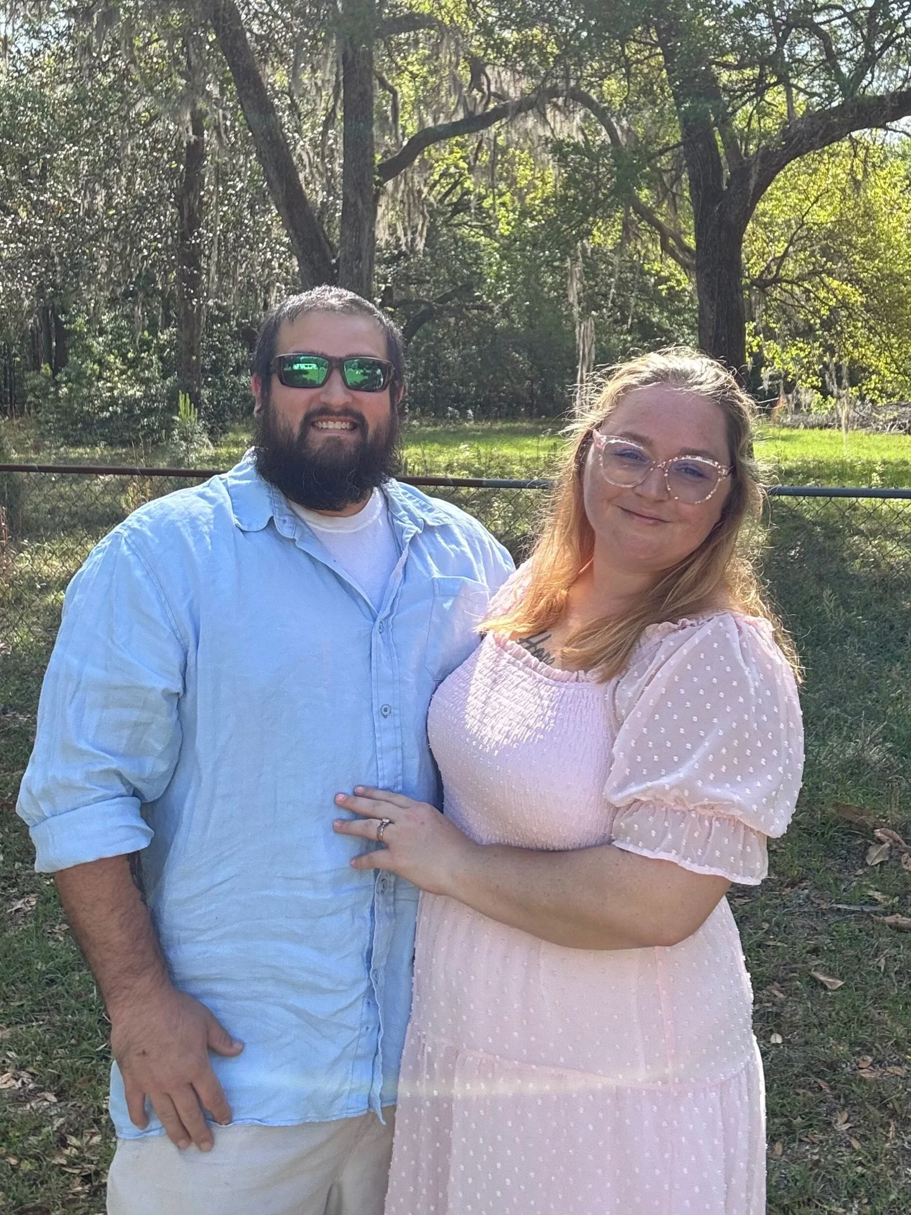 A couple standing outdoors in a green park with trees in the background, smiling at the camera. The man has a beard, sunglasses, and is wearing a light blue shirt. The woman has long blonde hair, glasses, and is wearing a pink dress with polka dots.