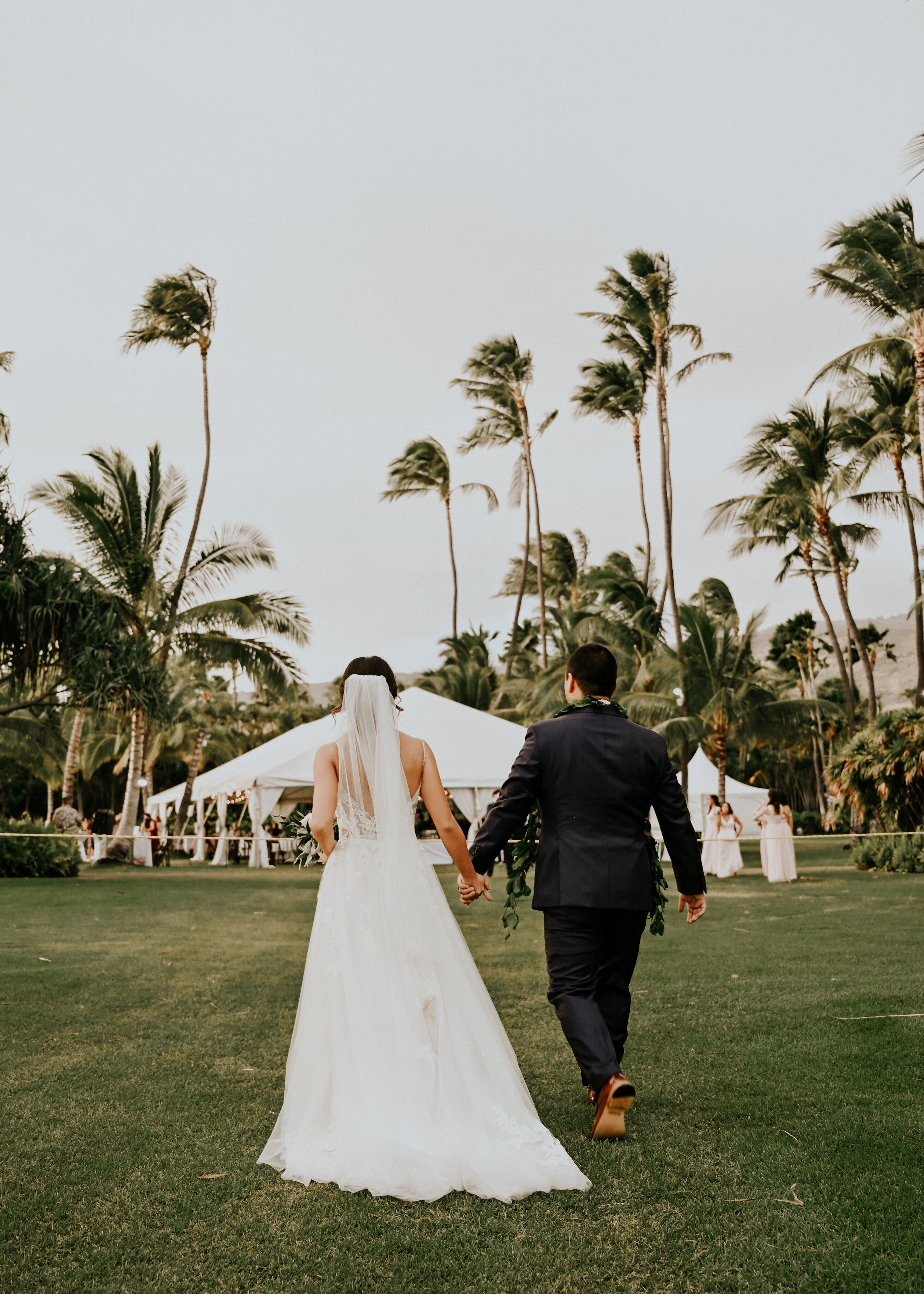 A bride and groom holding hands and walking on a golf course surrounded by palm trees in Hawaii, during a wedding ceremony outdoor in the evening. | Best Hawaii Wedding Photographer & Videographer | Oahu, Hawaii Island, Kauai, and Maui 