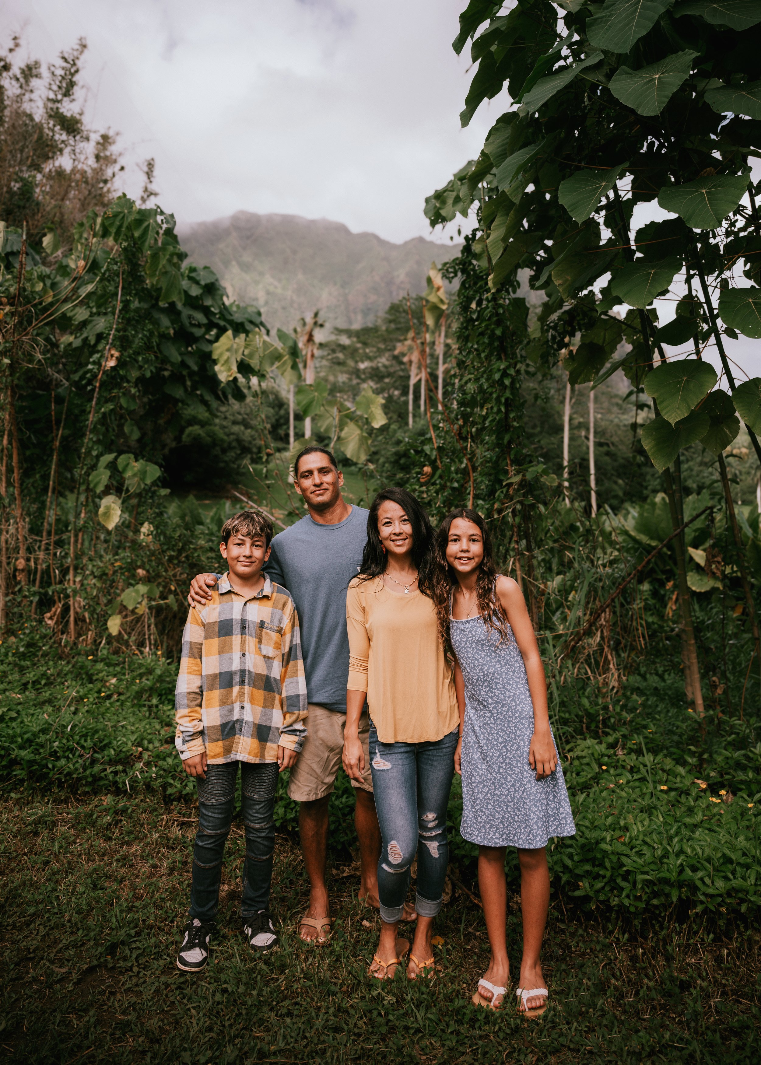 A family of five standing outdoors in a lush, green, mountainous area, smiling and posing for a photo. The group includes two young boys, a man, and two women, one of whom is wearing a yellow top and the other a gray dress. The background features large leafy trees and a mountain with some mist or clouds.