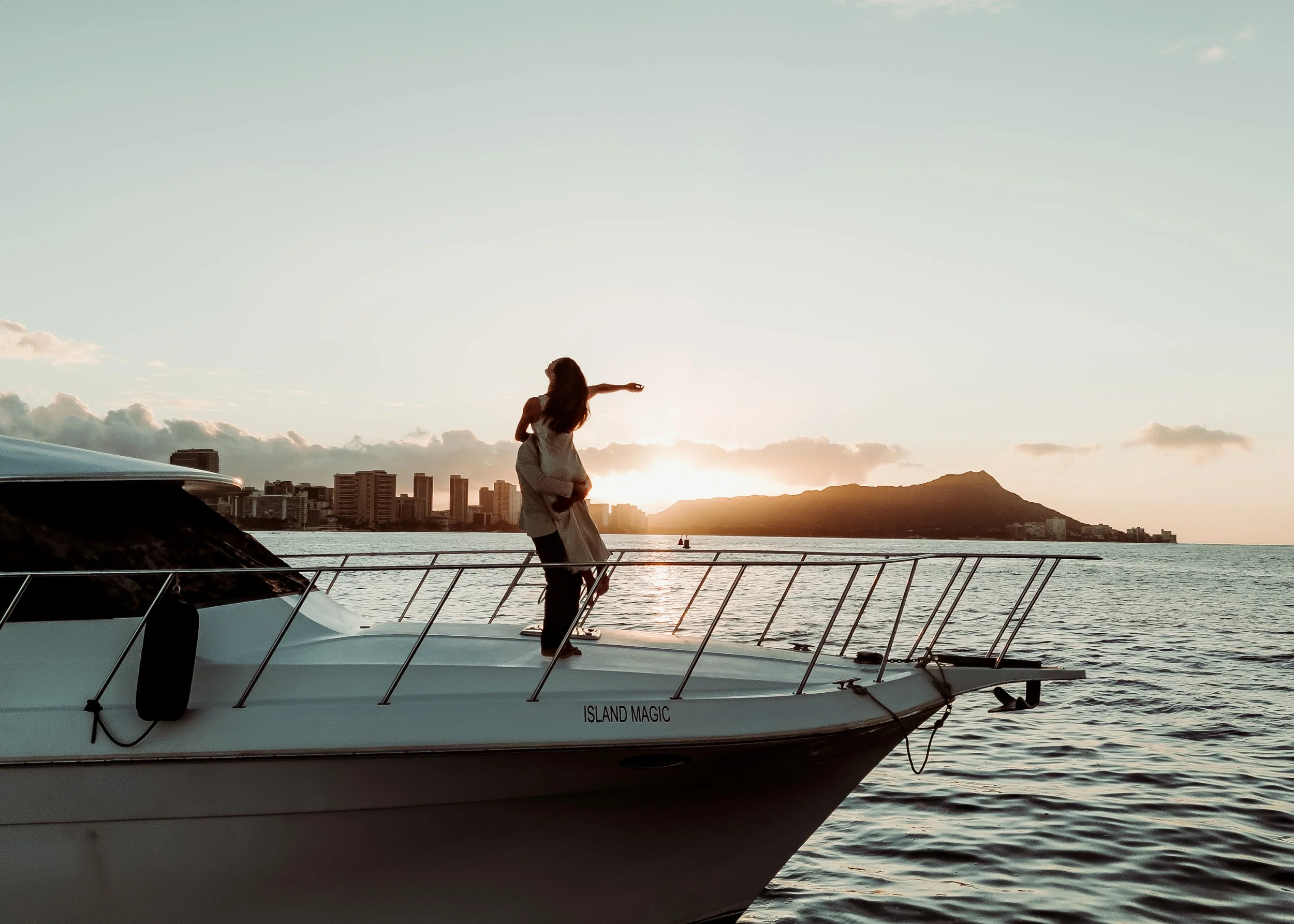 A couple standing on the deck of a yacht named Island Magic, with her arm outstretched towards the sunset over the city skyline and mountains in the background.  | Best Hawaii Wedding Photographer & Videographer | Oahu, Hawaii Island, Kauai, and Maui