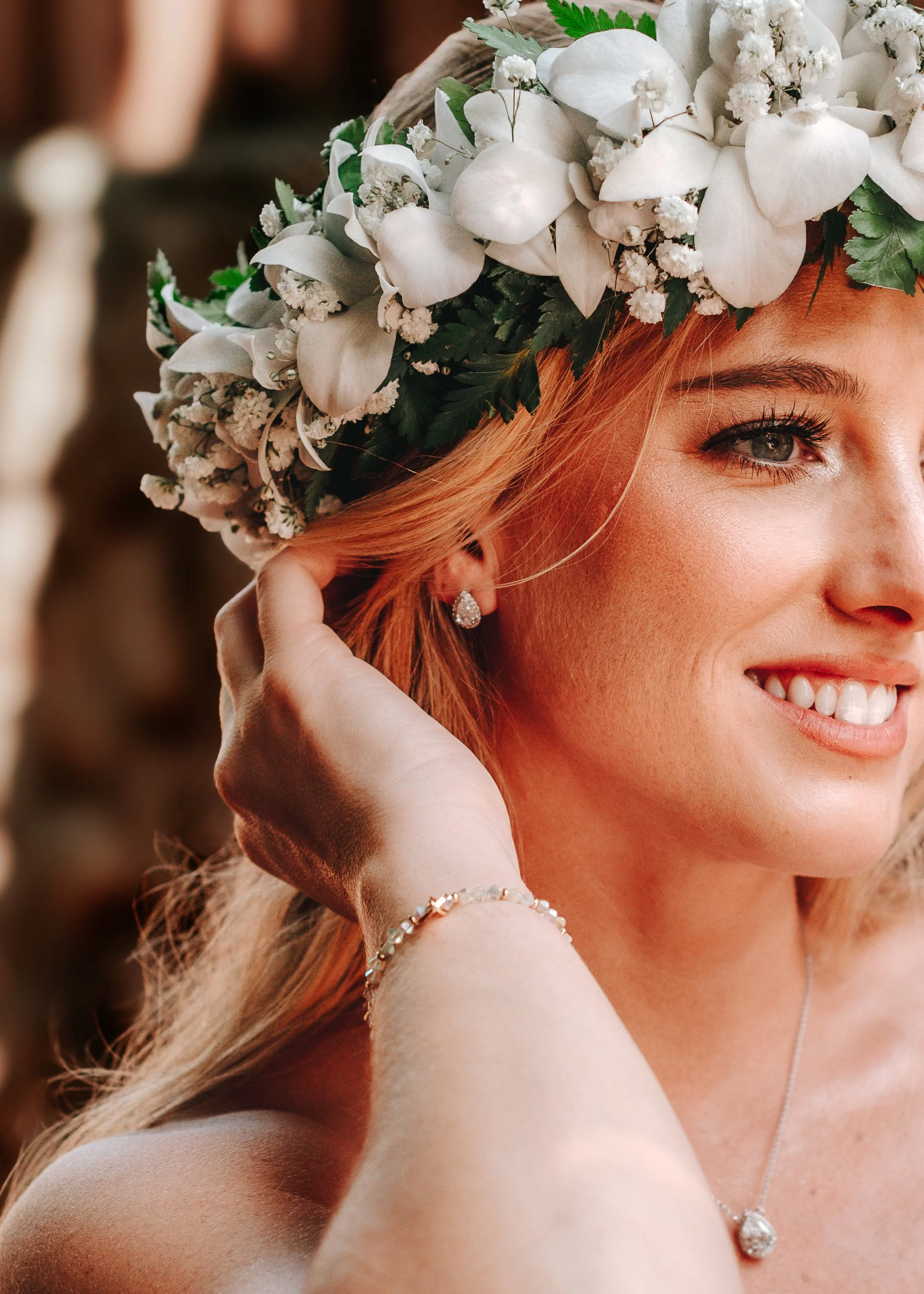 A woman with blonde hair wearing a floral crown made of white flowers and greenery, smiling, showing her teeth, and wearing jewelry including a pearl stud earring, a bracelet, and a necklace with a teardrop-shaped pendant.