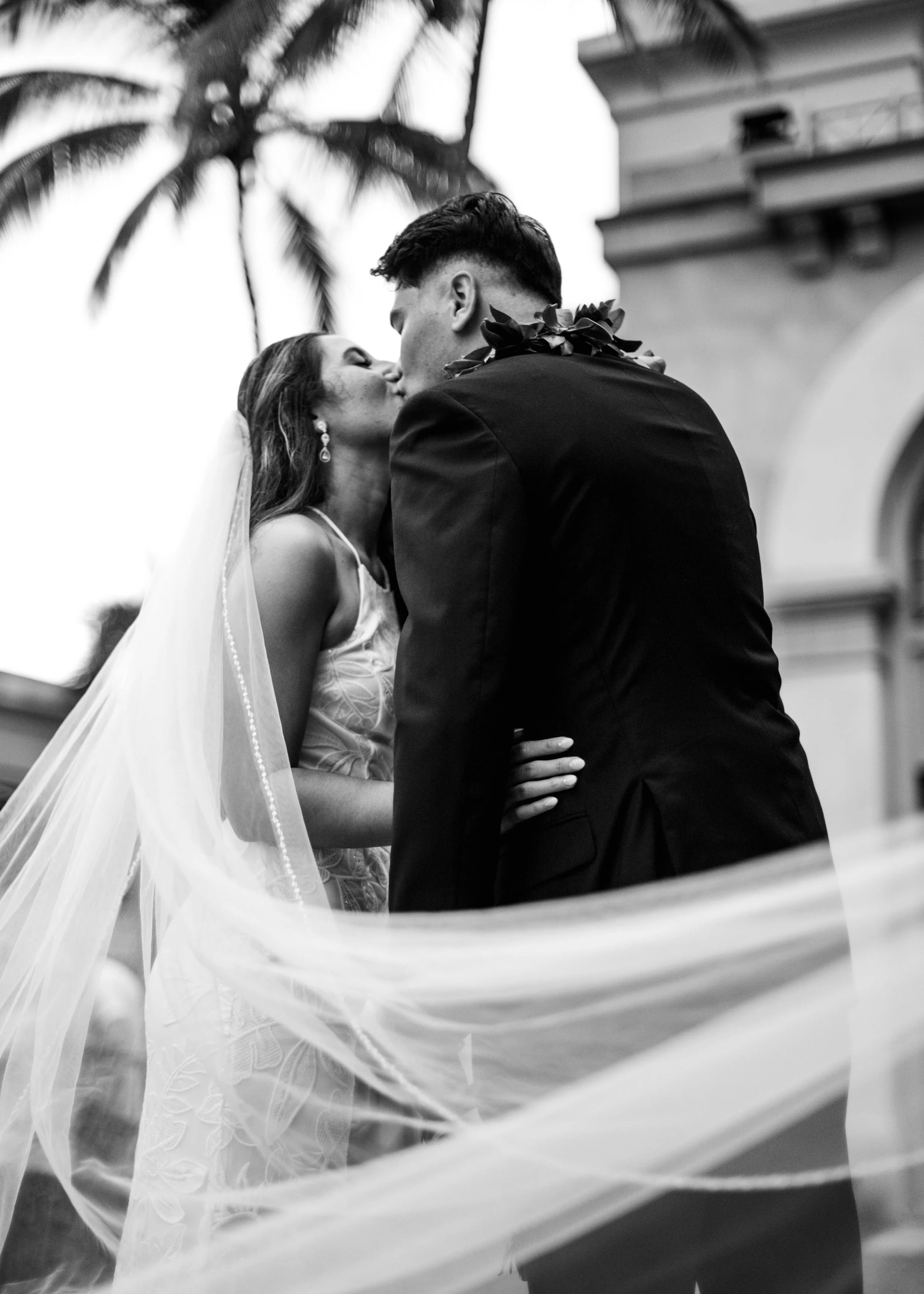 A black and white photo of a couple sharing a kiss outdoors, with the woman wearing a wedding dress and veil, and the man dressed in a suit with a floral lei around his neck, in front of a building and tall palm trees. | Best Hawaii Wedding Photograp