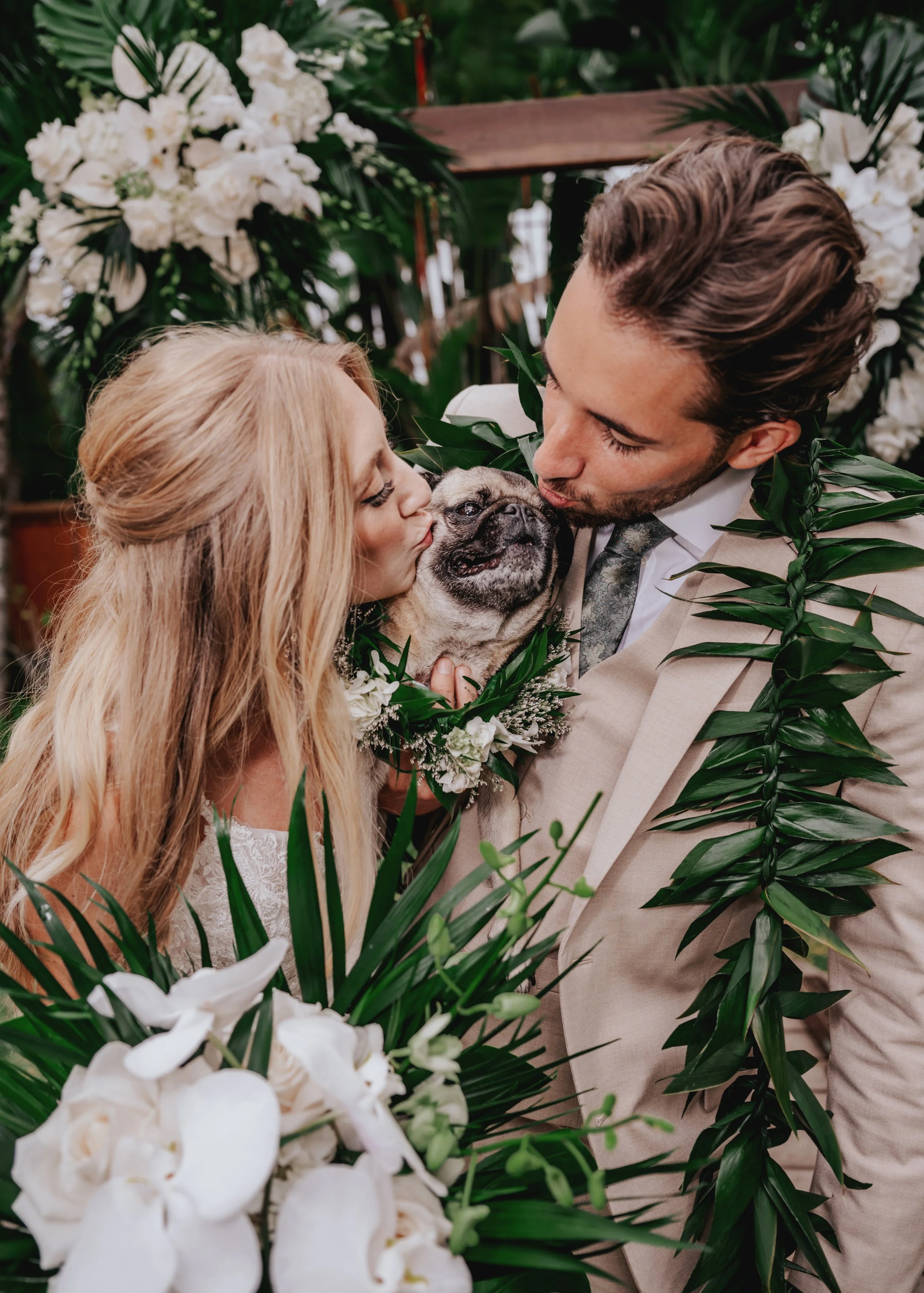 A couple kisses their wedding dog, a pug, surrounded by white flowers and green foliage at their wedding ceremony. | Best Hawaii Wedding Photographer & Videographer | Oahu, Hawaii Island, Kauai, and Maui 
