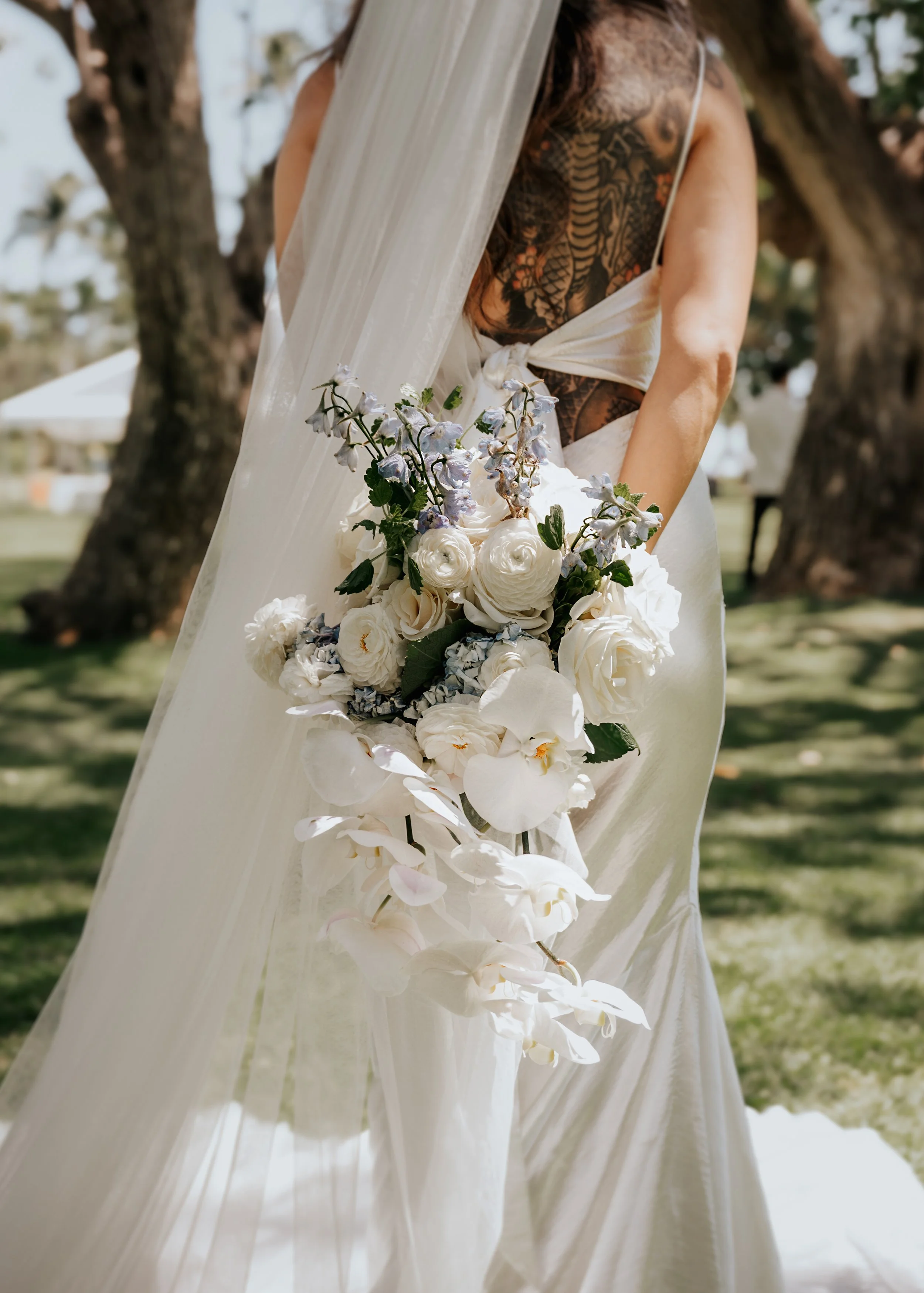 A bride holding a bouquet of white and pale purple flowers outdoors, with trees and a white tent in the background.  | Best Hawaii Wedding Photographer & Videographer | Oahu, Hawaii Island, Kauai, and Maui