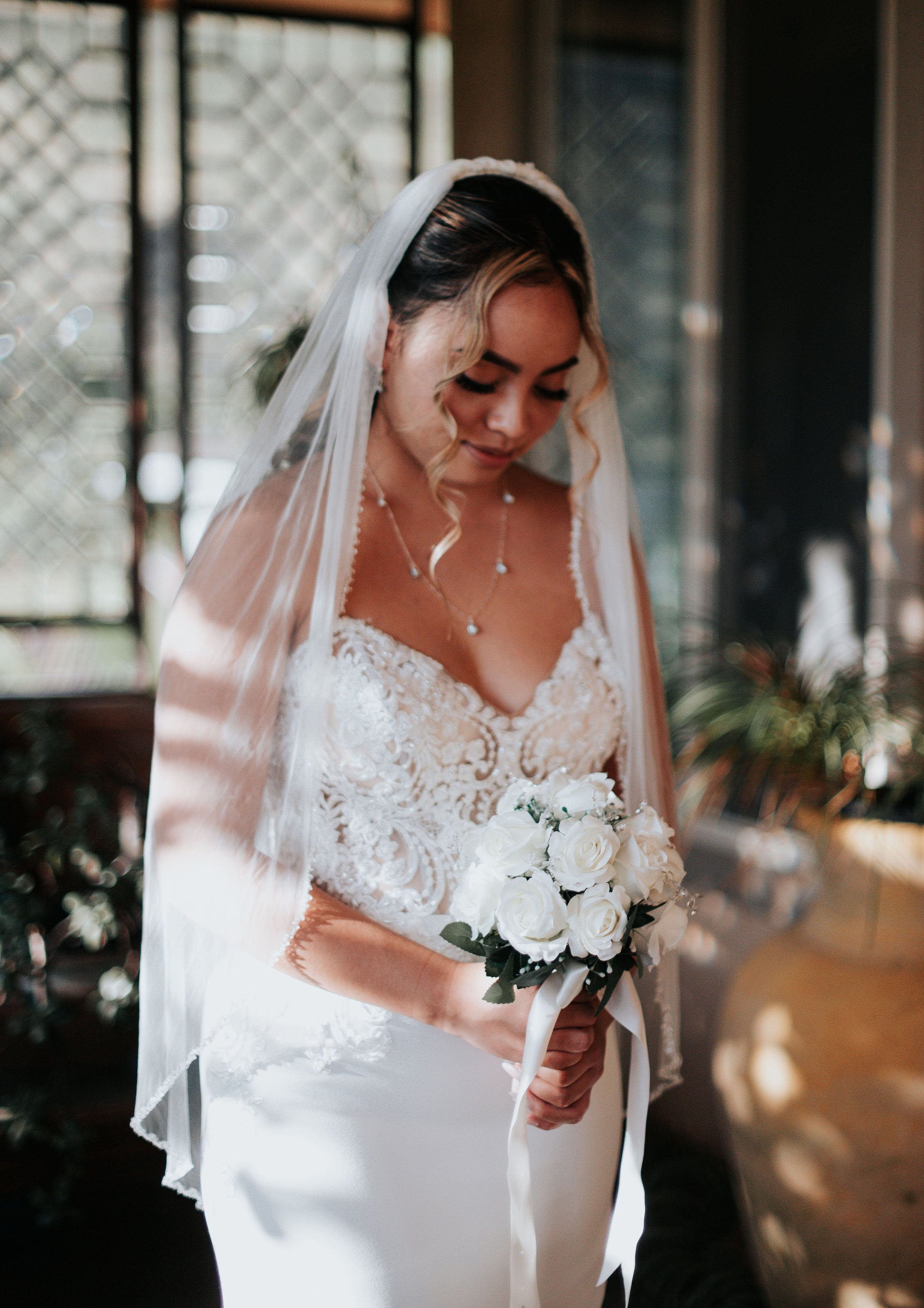 A bride in a white lace wedding dress holding a bouquet of white roses, wearing a veil and jewelry, inside a warmly lit indoor space. | Best Hawaii Wedding Photographer & Videographer | Oahu, Hawaii Island, Kauai, and Maui 