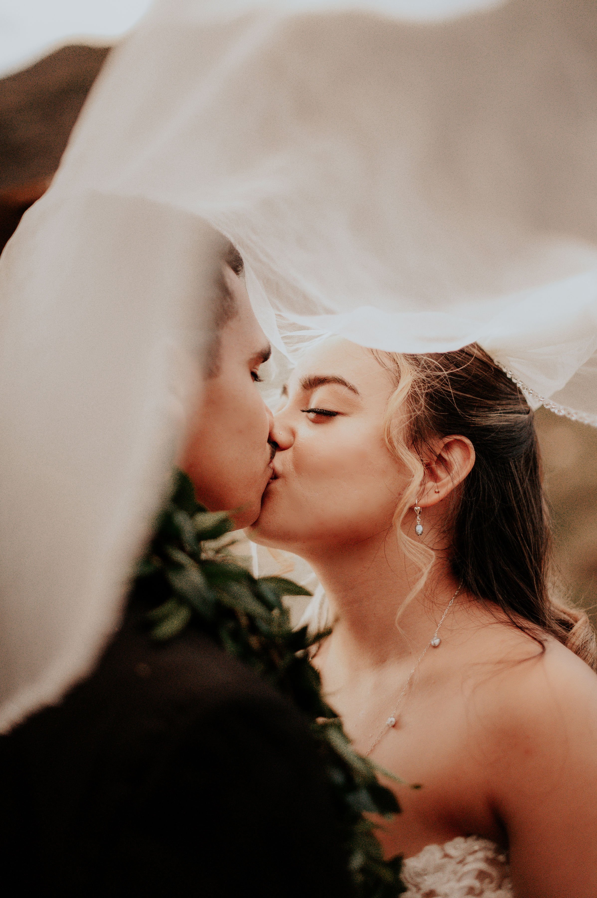 A newlywed couple sharing a kiss during their wedding, with partially visible bride in a veil and earrings, and groom in a tuxedo, in a closely focused shot.  | Best Hawaii Wedding Photographer & Videographer | Oahu, Hawaii Island, Kauai, and Maui