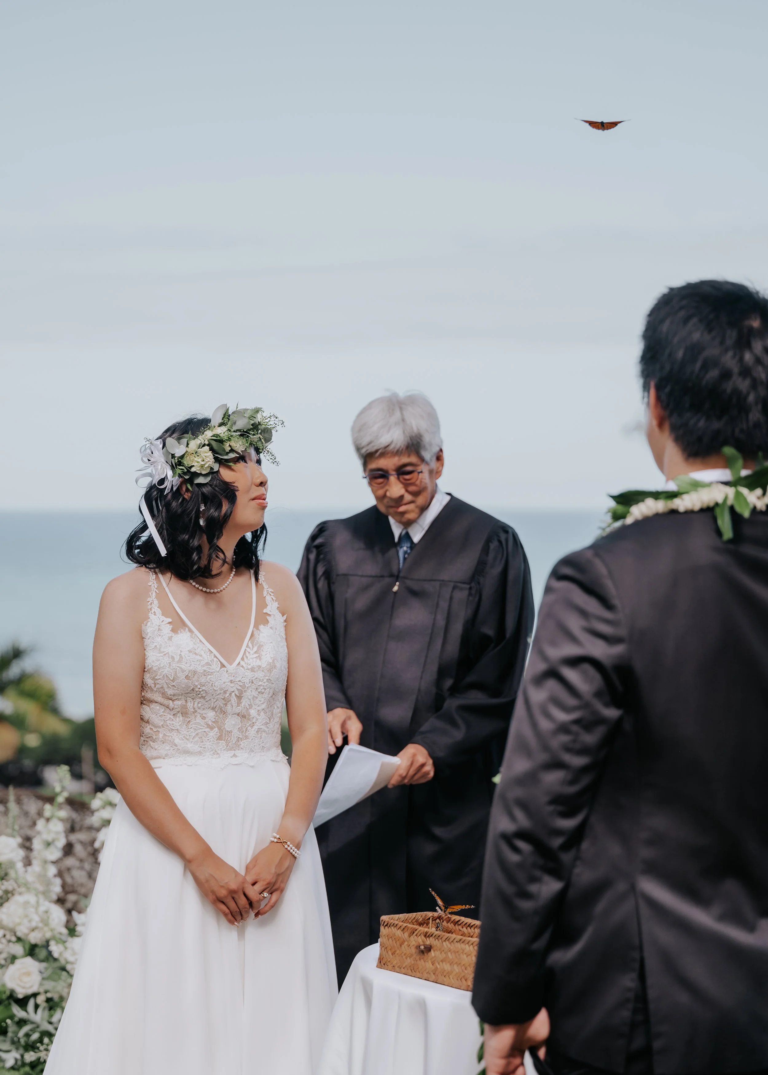 A couple is getting married outdoors with an officiant, with the ocean in the background. The bride is wearing a white wedding dress with lace details and a floral crown, and the groom and another man are dressed in suits. A butterfly is flying in th