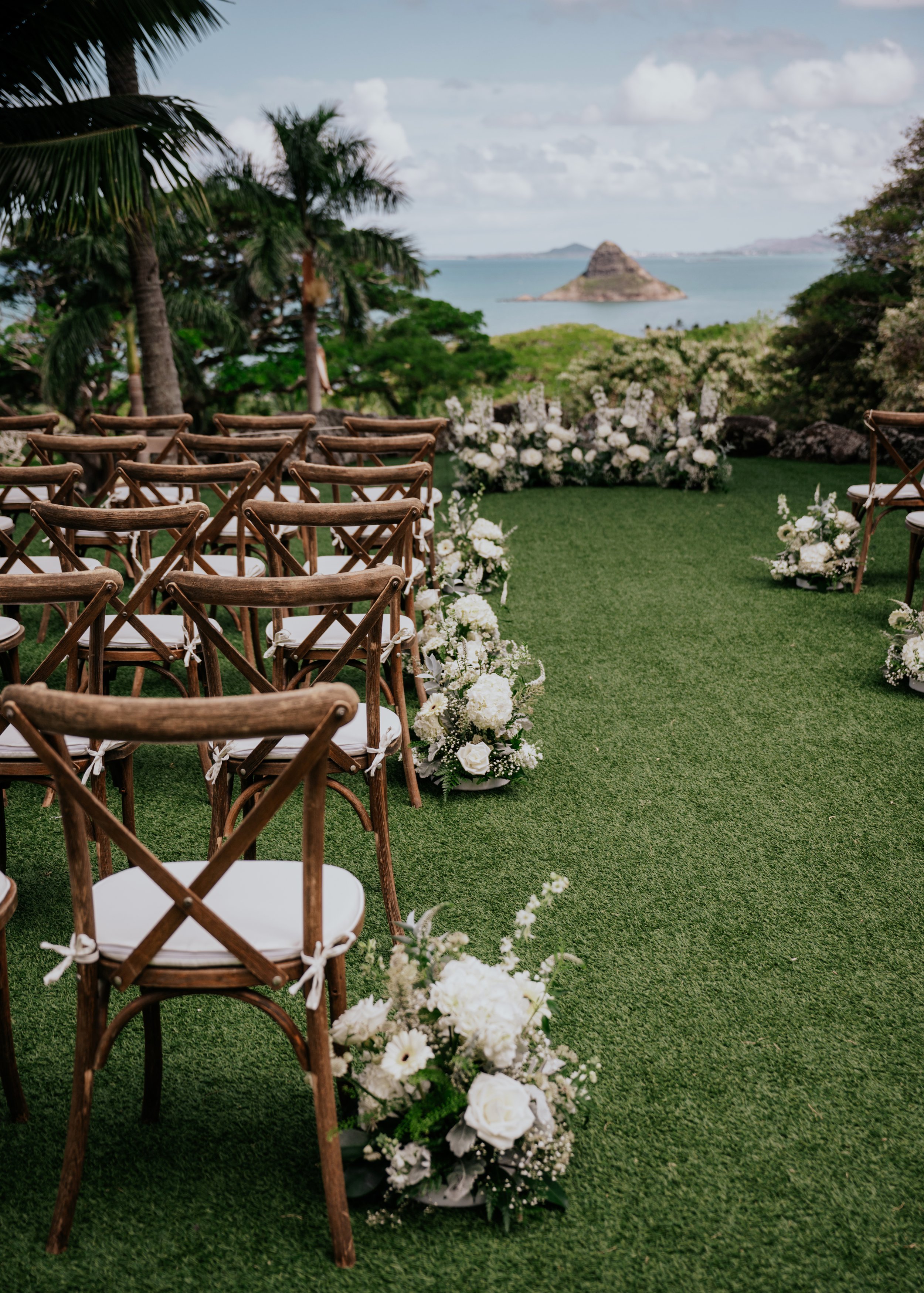 Outdoor wedding setup in Oahu with wooden chairs arranged on green grass, decorated with white flowers along the aisle, overlooking a scenic view of the ocean with a small island in the distance under a cloudy sky.