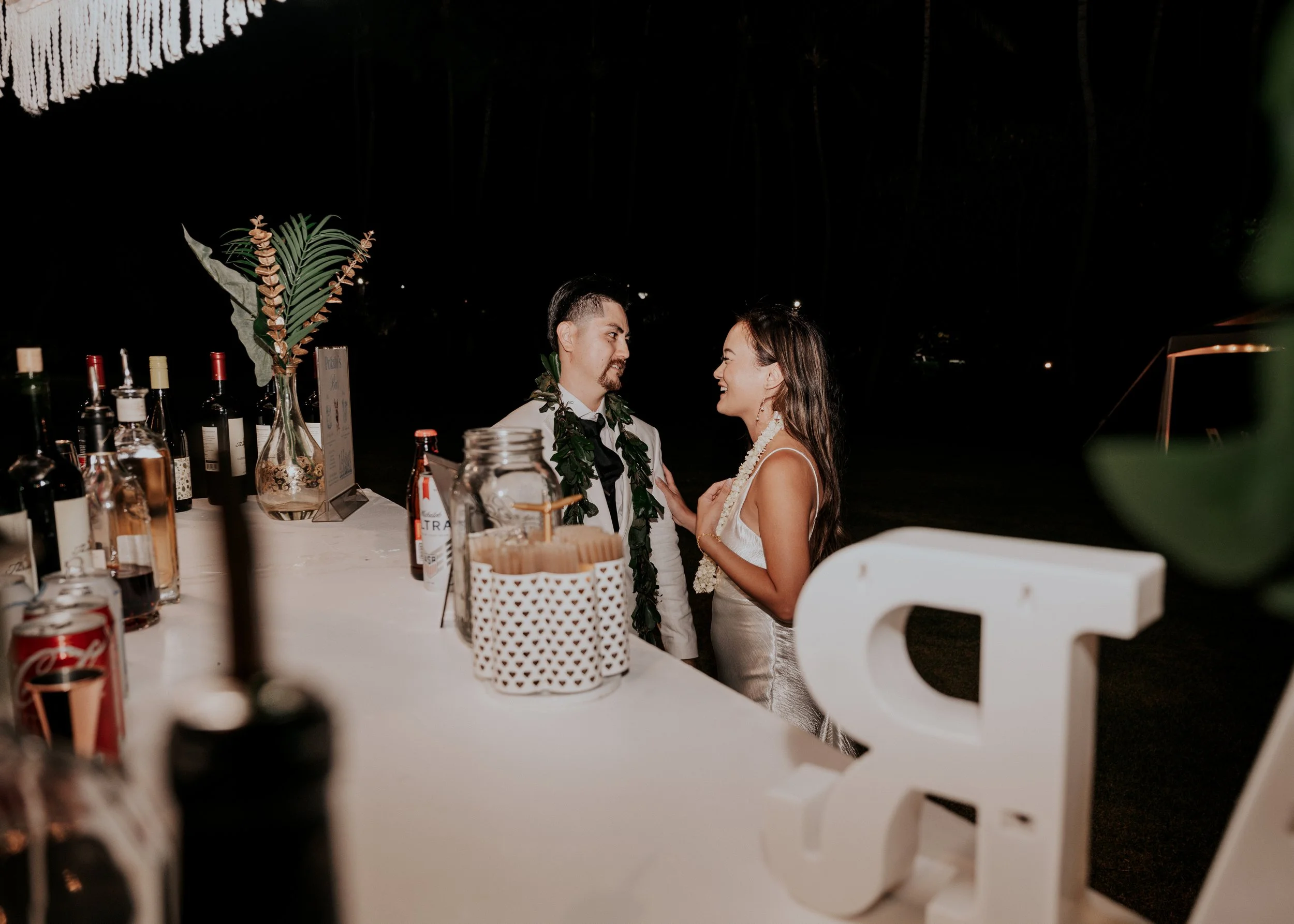 A couple dressed in wedding attire sharing a happy moment at the bar during their reception, with a table decorated with bottles, glasses, and a large tropical leaf in the foreground.  | Best Hawaii Wedding Photographer & Videographer | Oahu, Hawaii