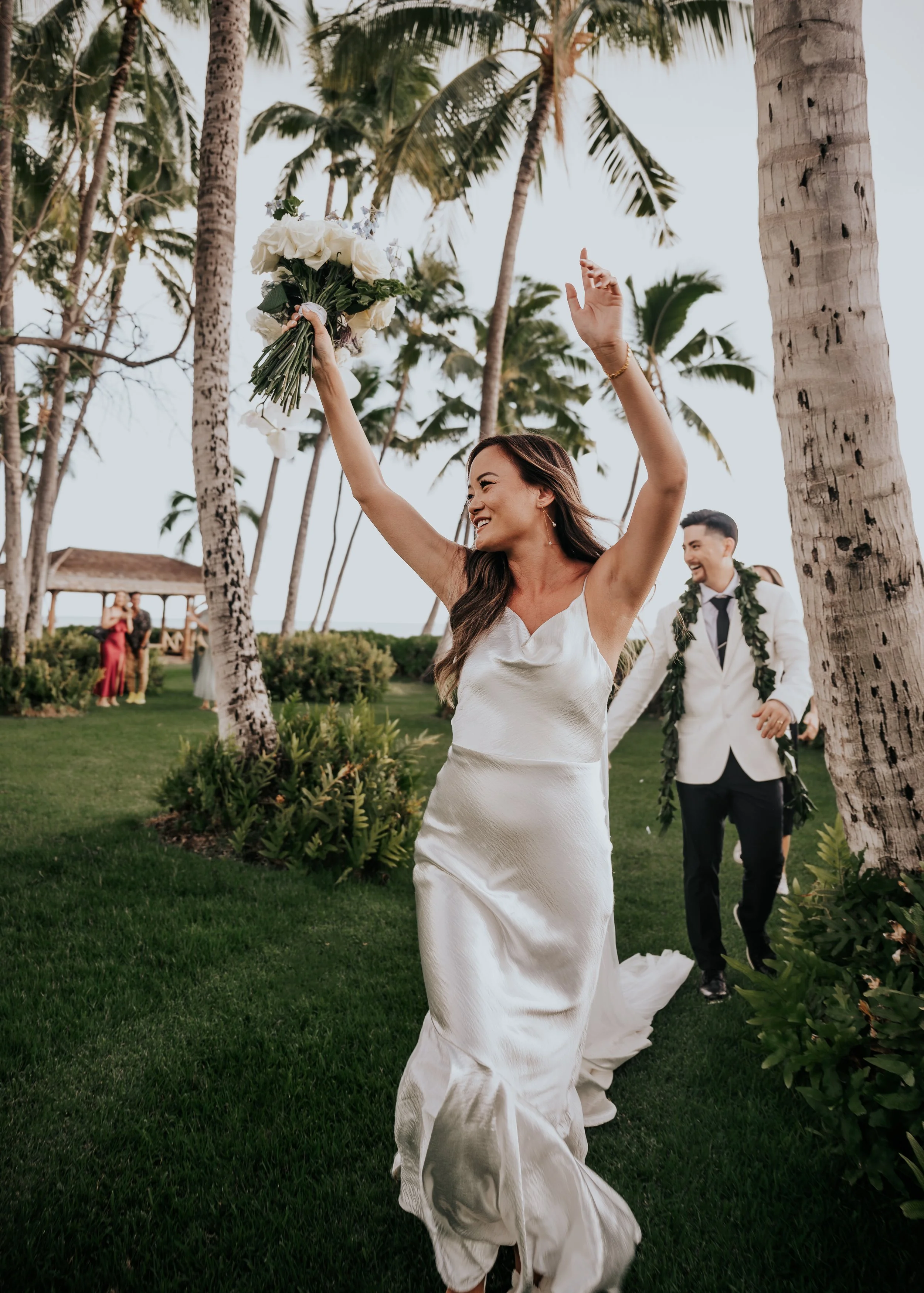A bride in a white wedding dress holding a bouquet and celebrating outdoors with palm trees in the background.  | Best Hawaii Wedding Photographer & Videographer | Oahu, Hawaii Island, Kauai, and Maui