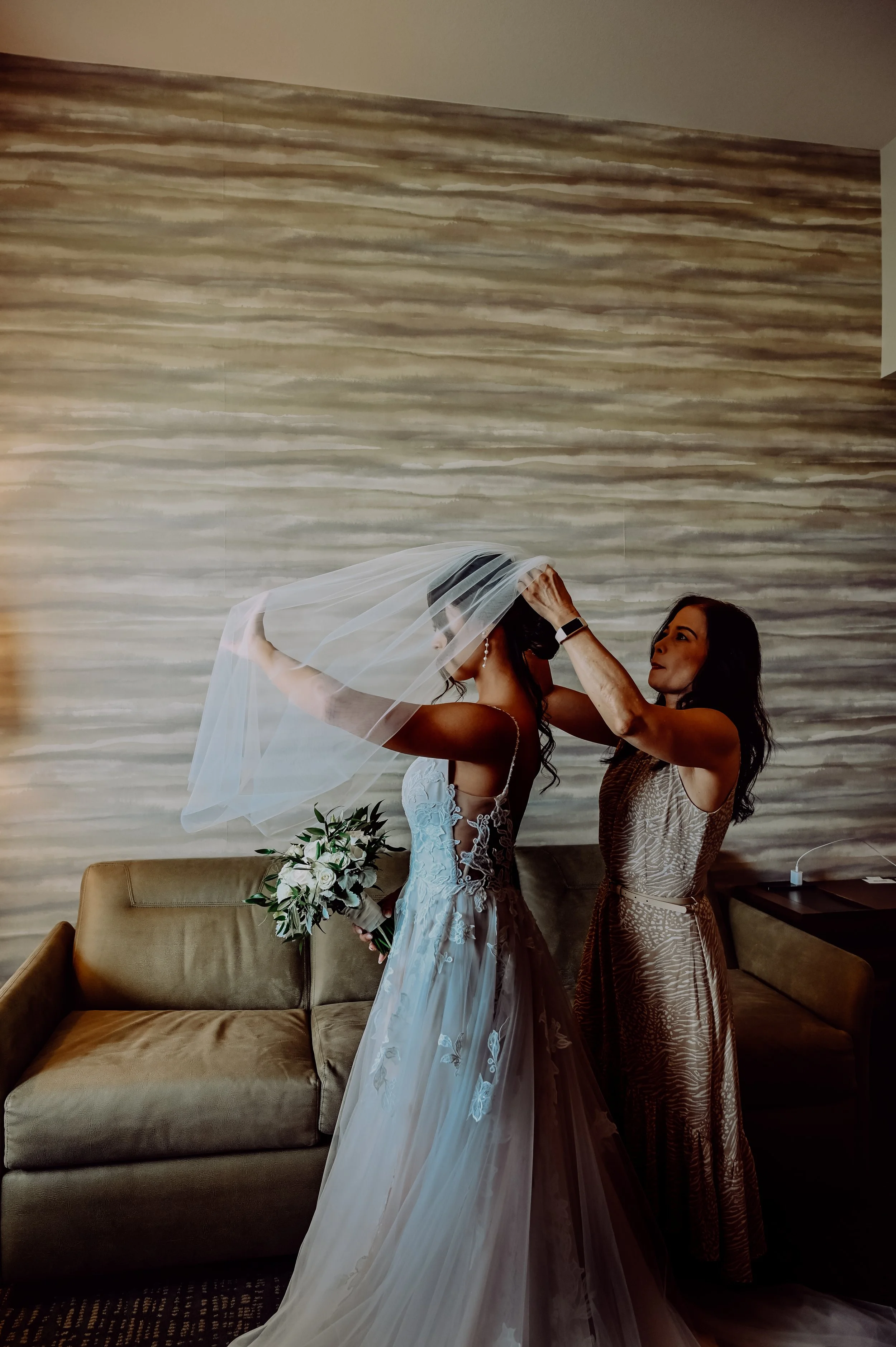 A bride in a white wedding gown holding a bouquet, while a woman helps her put on a veil in a room with a brown and beige striped wall.  | Best Hawaii Wedding Photographer & Videographer | Oahu, Hawaii Island, Kauai, and Maui