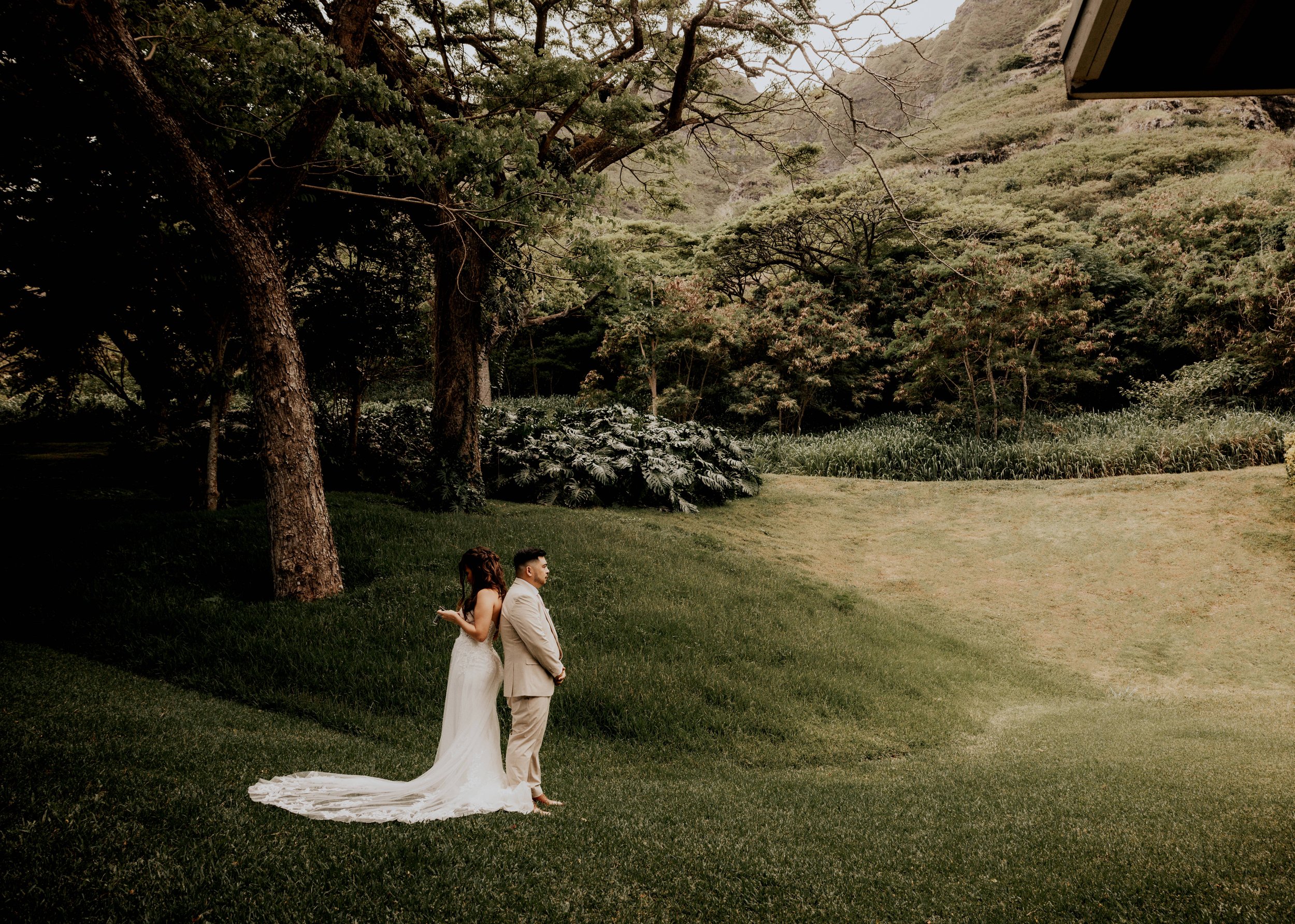 A bride and groom standing back to back on a grassy area outdoors during their first look with the bride in a white wedding dress and the groom in a light-colored suit, surrounded by trees and greenery reciting their vows.  | Photographer & Video