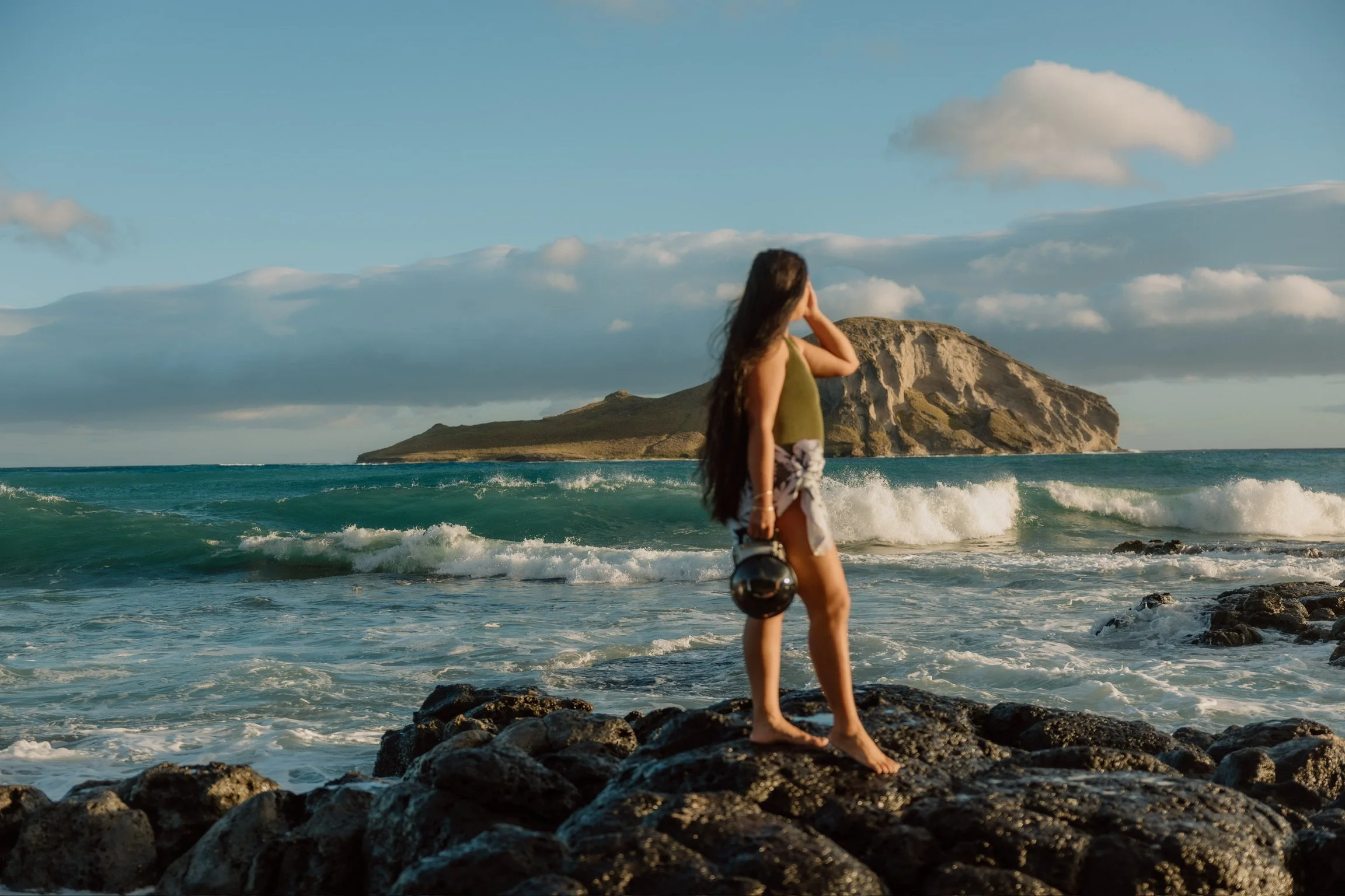 A woman, the owner, Ashley Gallarde with long dark hair standing on rocks at the beach, holding a water housing camera facing the ocean with an island and cloudy sky in the background.