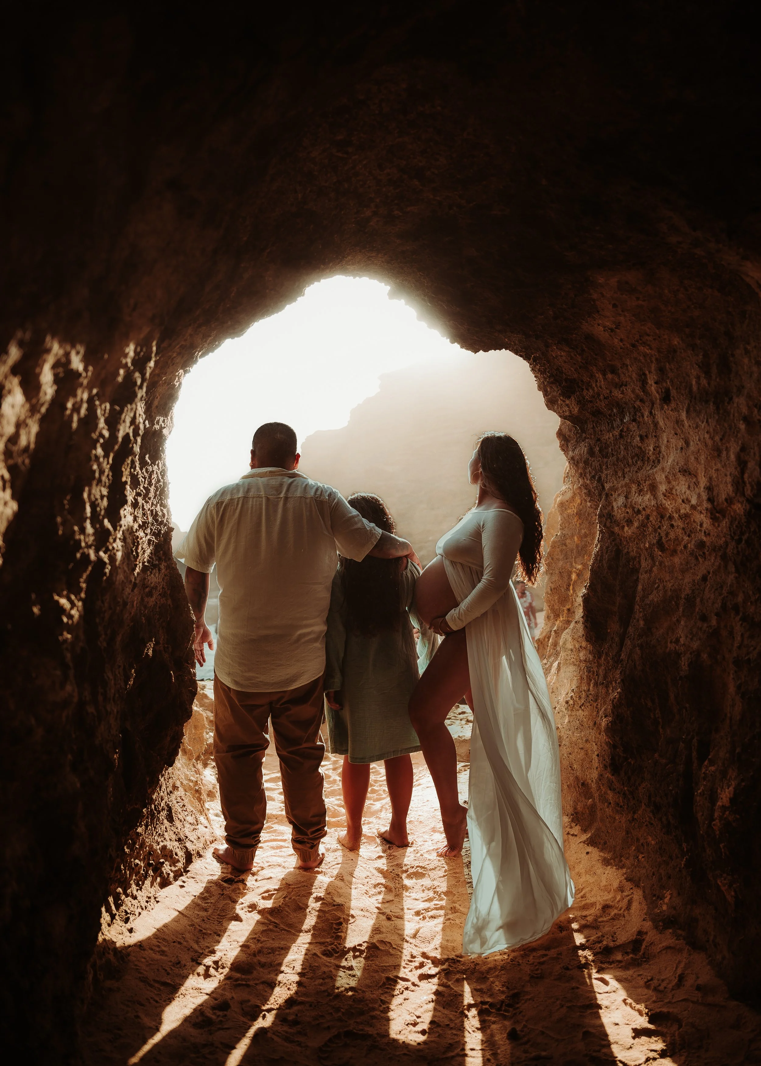 A group of five people, including a pregnant woman, standing in a rocky cave opening, with sunlight shining behind them, casting long shadows on the sandy ground.
