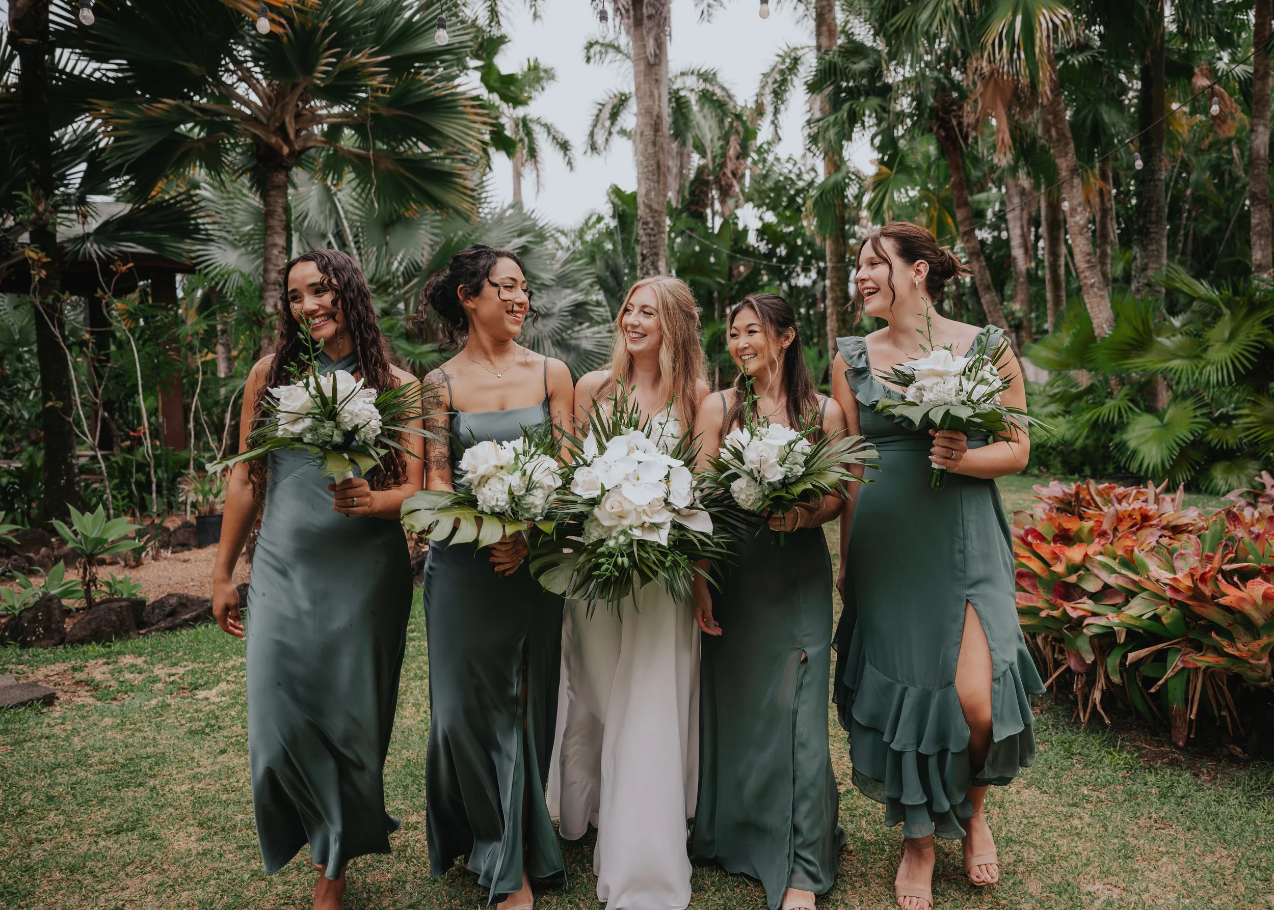 a bridal party of five women dressed in matching satin dresses holding bouquets, walking through a lush garden with palm trees, smiling and laughing.  | Best Hawaii Wedding Photographer & Videographer | Oahu, Hawaii Island, Kauai, and Maui