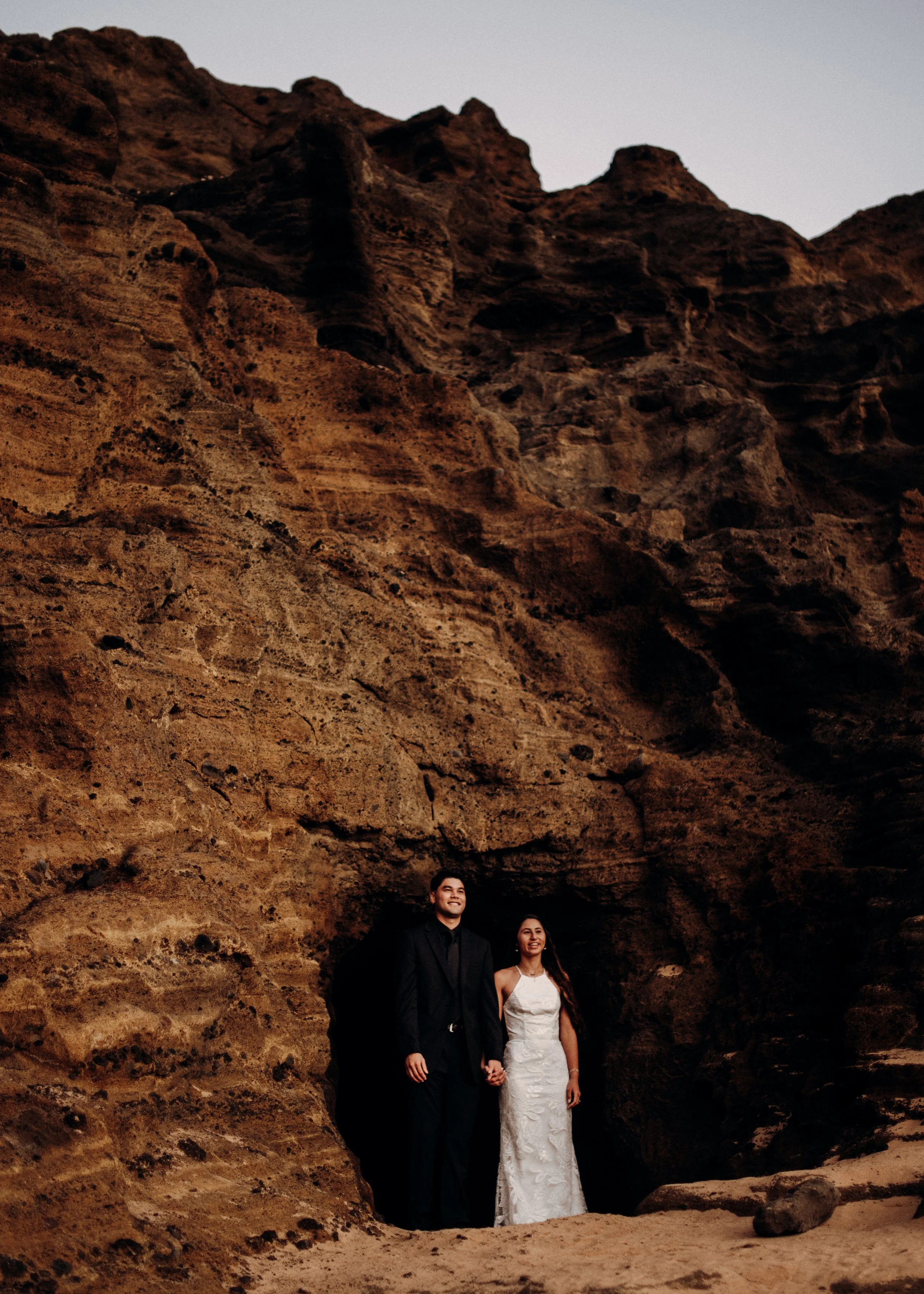 A newlywed couple standing hand in hand in front of a large rocky canyon that is a lava tube in hawaii during sunrise.  | Best Hawaii Wedding Photographer & Videographer | Oahu, Hawaii Island, Kauai, and Maui