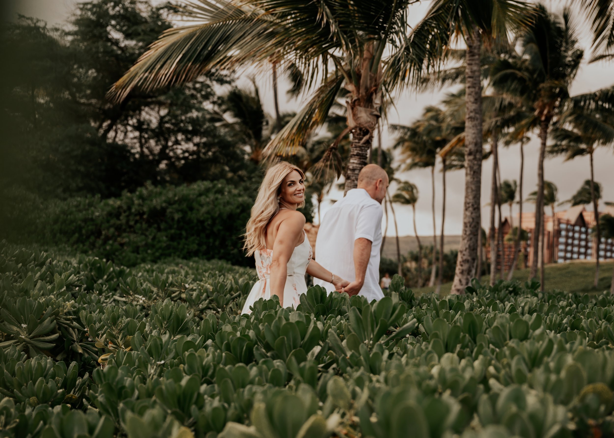 A smiling woman with long blond hair in a white dress holding hands with a man in a white shirt as they walk through a lush, tropical garden with palm trees and green foliage.