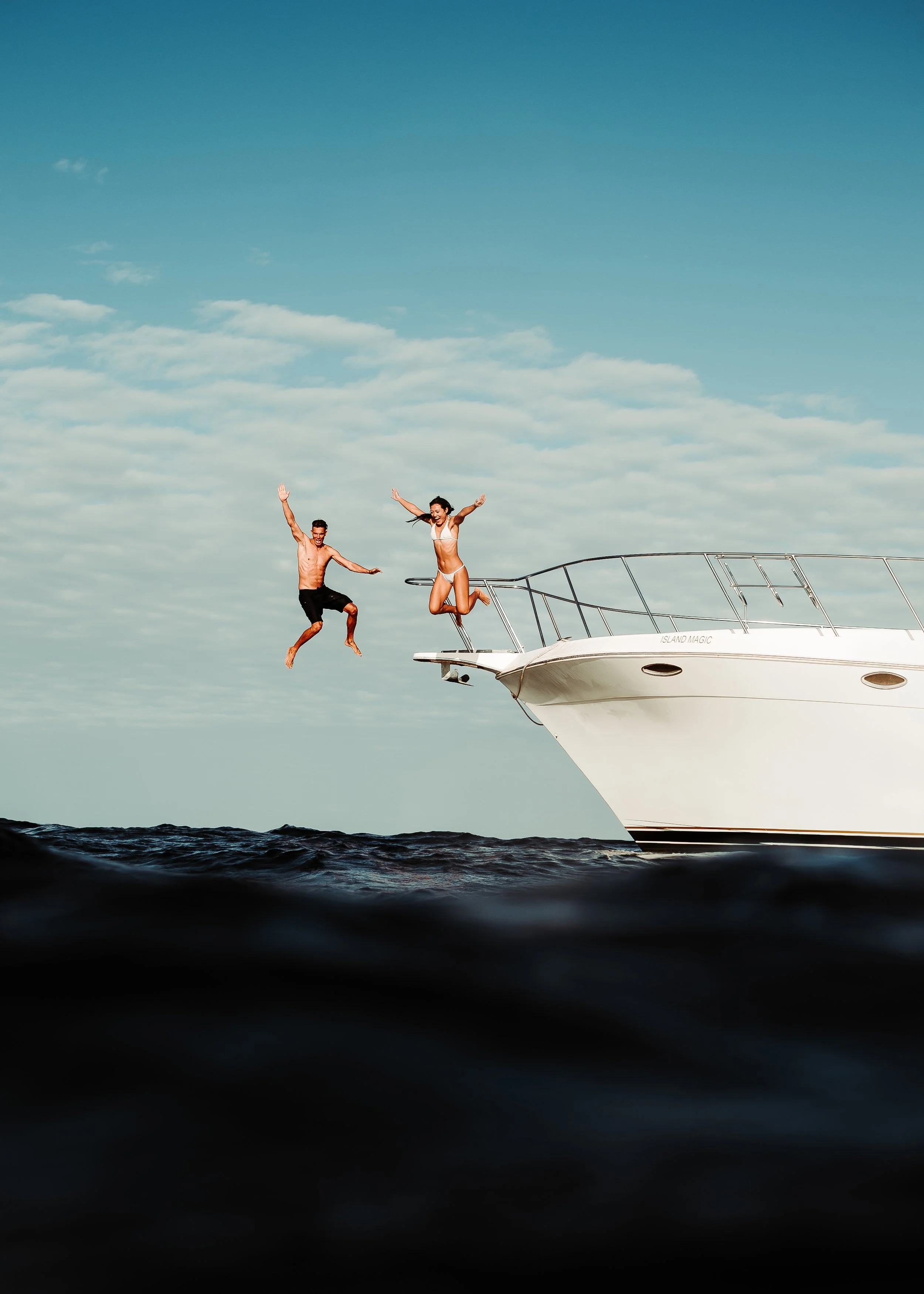 Two people jump into the water from the side of a large white yacht named 'Island Magic' on a cloudy day.