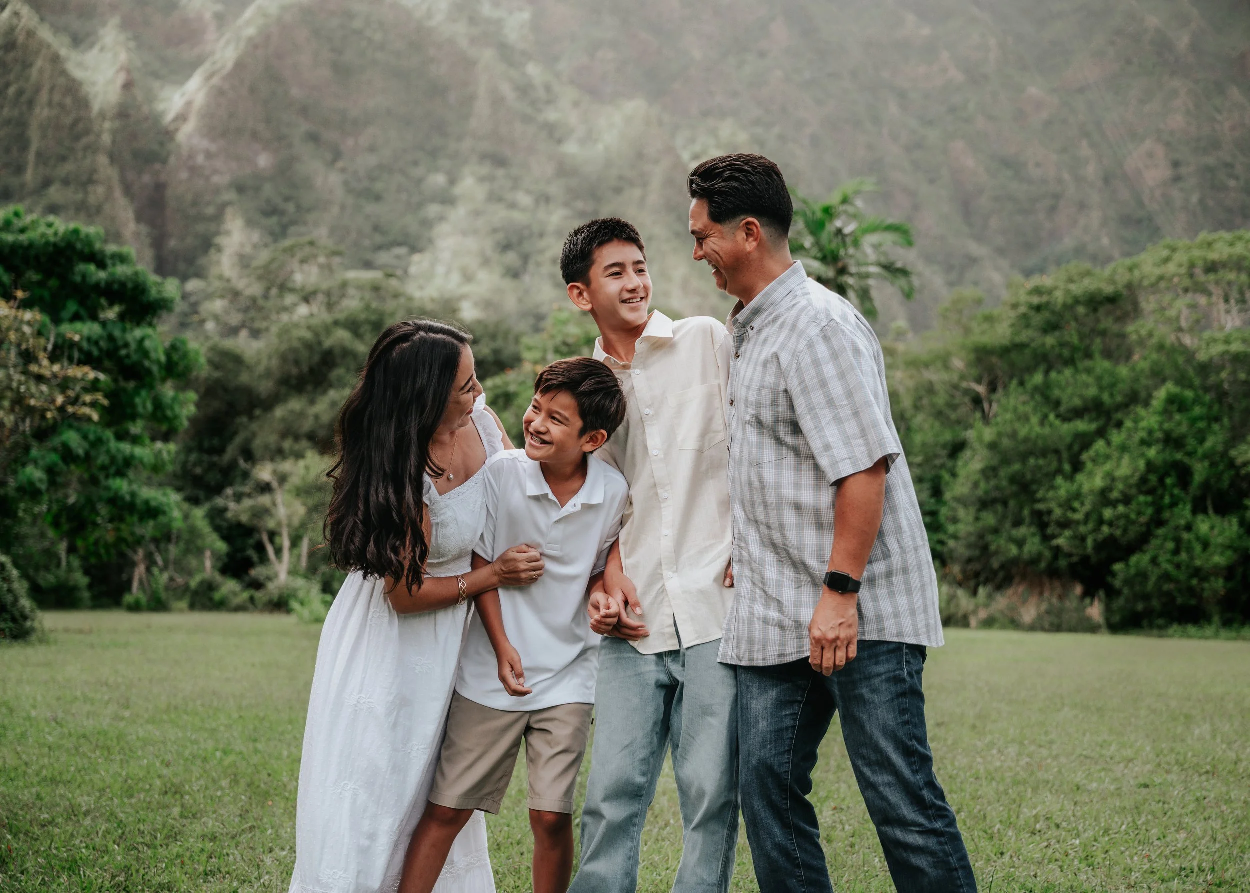 A family of five smiling and laughing outdoors in a grassy area with lush green trees and mountains in the background.