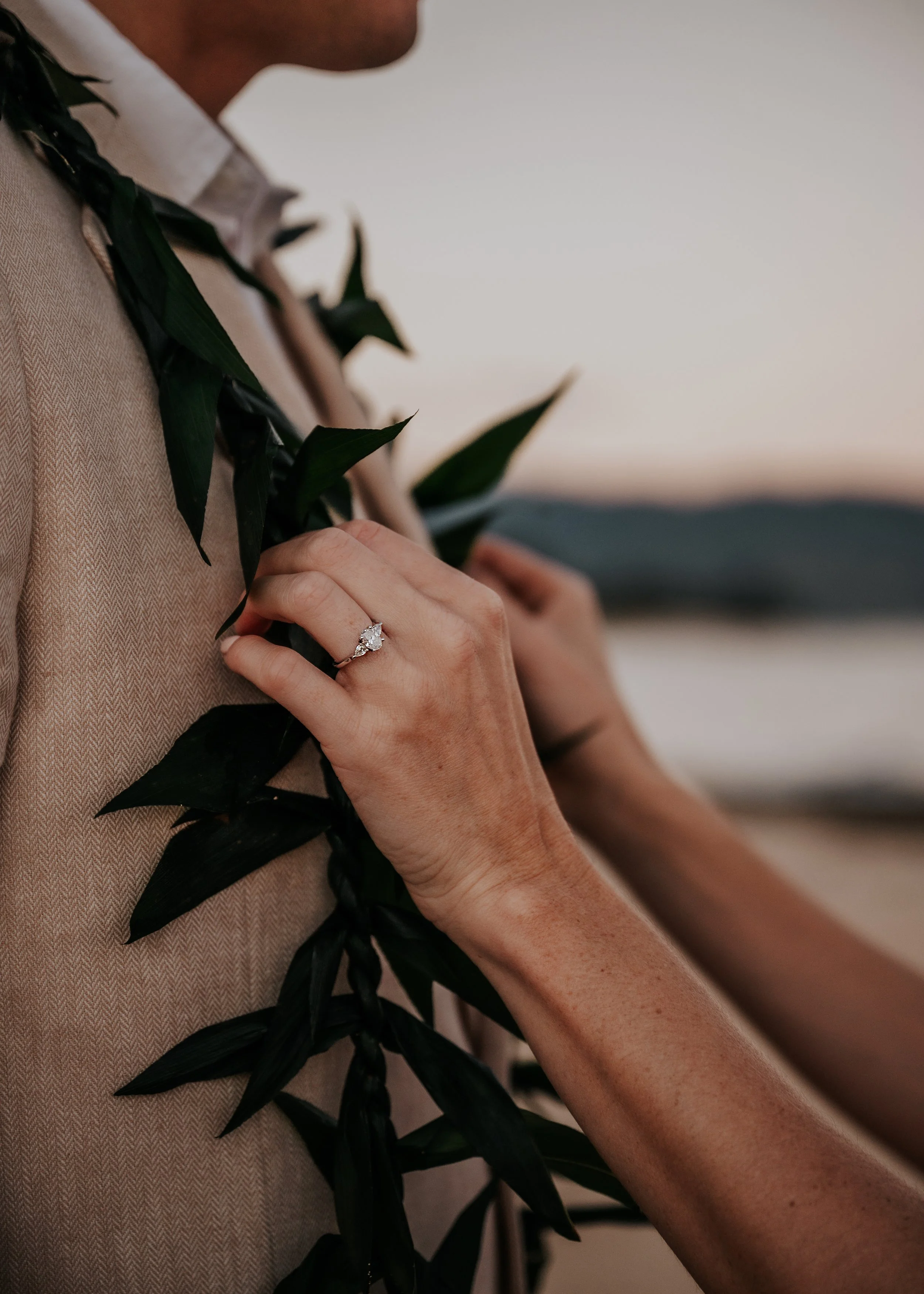 A person wearing a beige outfit with a white shirt and a couple of rings on their finger, holding green leaves close to their chest.