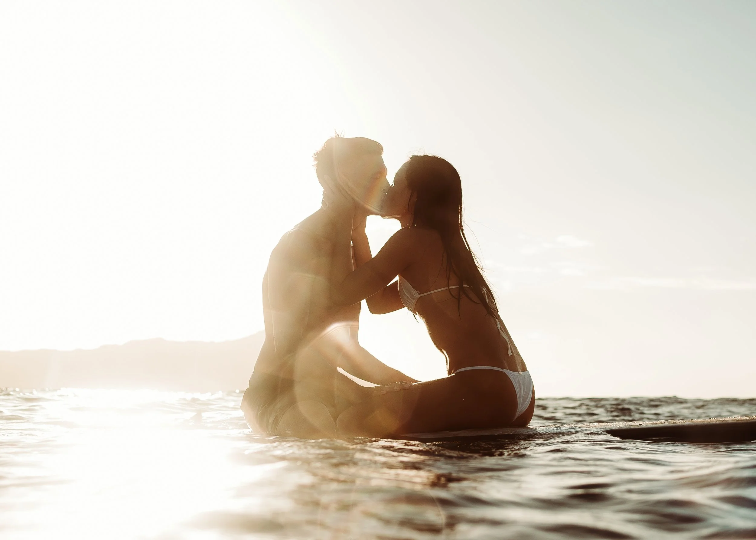 A couple sitting on a surfboard in the water kissing, with the sun rising behind them.  | Best Hawaii Wedding Photographer & Videographer | Oahu, Hawaii Island, Kauai, and Maui
