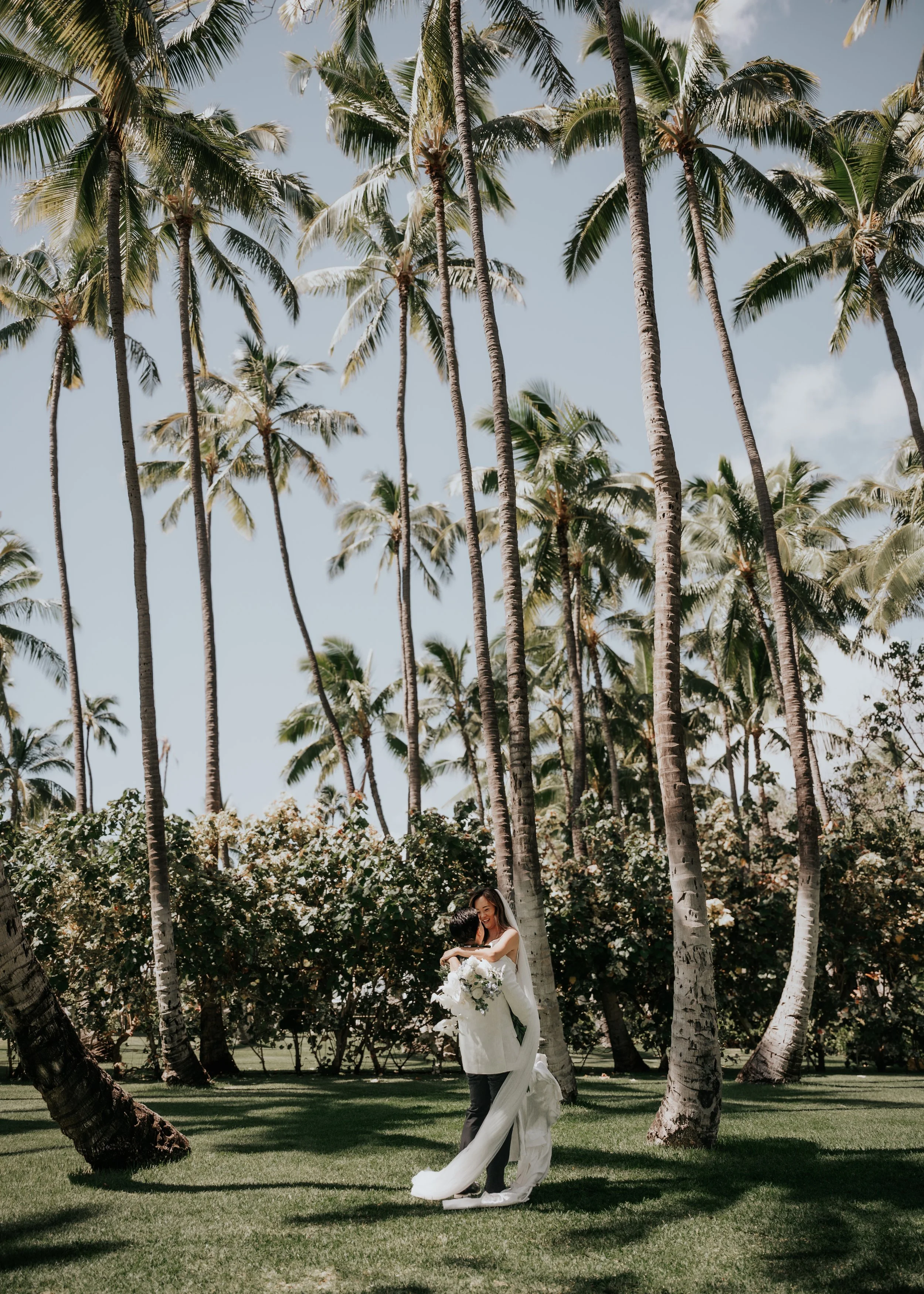 Newlyweds stands in a lush outdoor setting with tall palm trees. The woman wears a white wedding gown and the man in dark pants and a white shirt. They appear to be smiling and in a joyful moment, with the woman holding a bouquet of flowers.