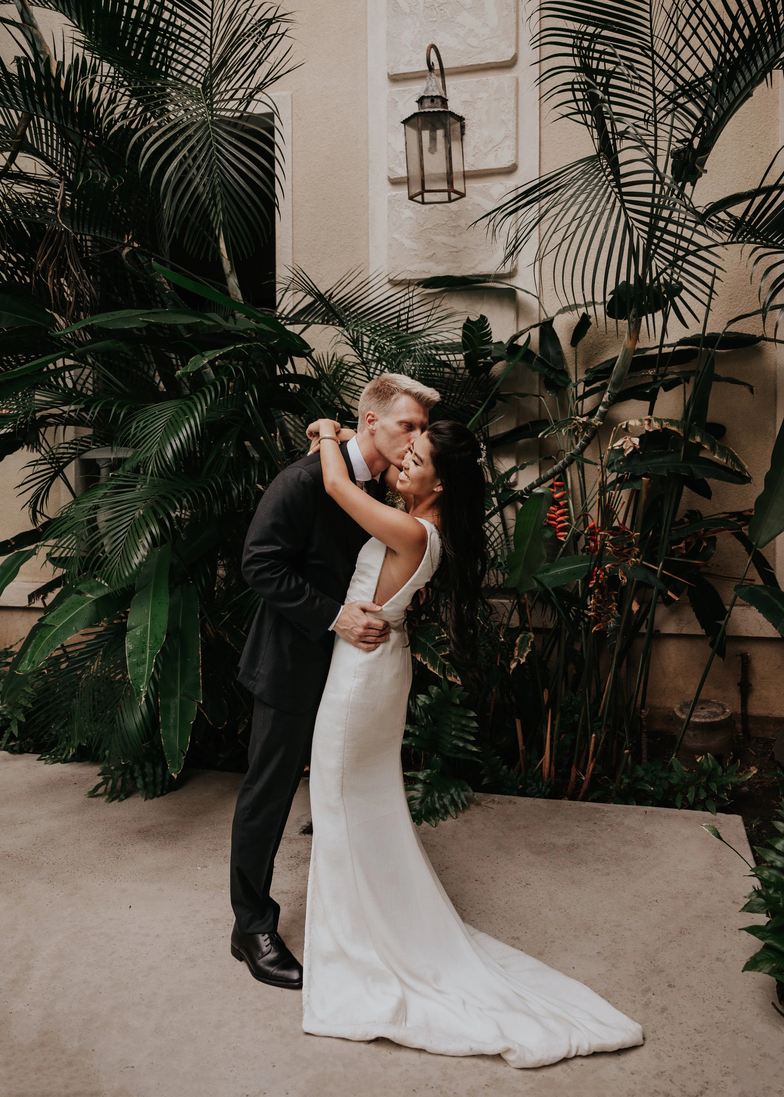 A bride and groom in wedding attire sharing a kiss and embracing each other in a lush outdoor garden setting surrounded by tropical plants.