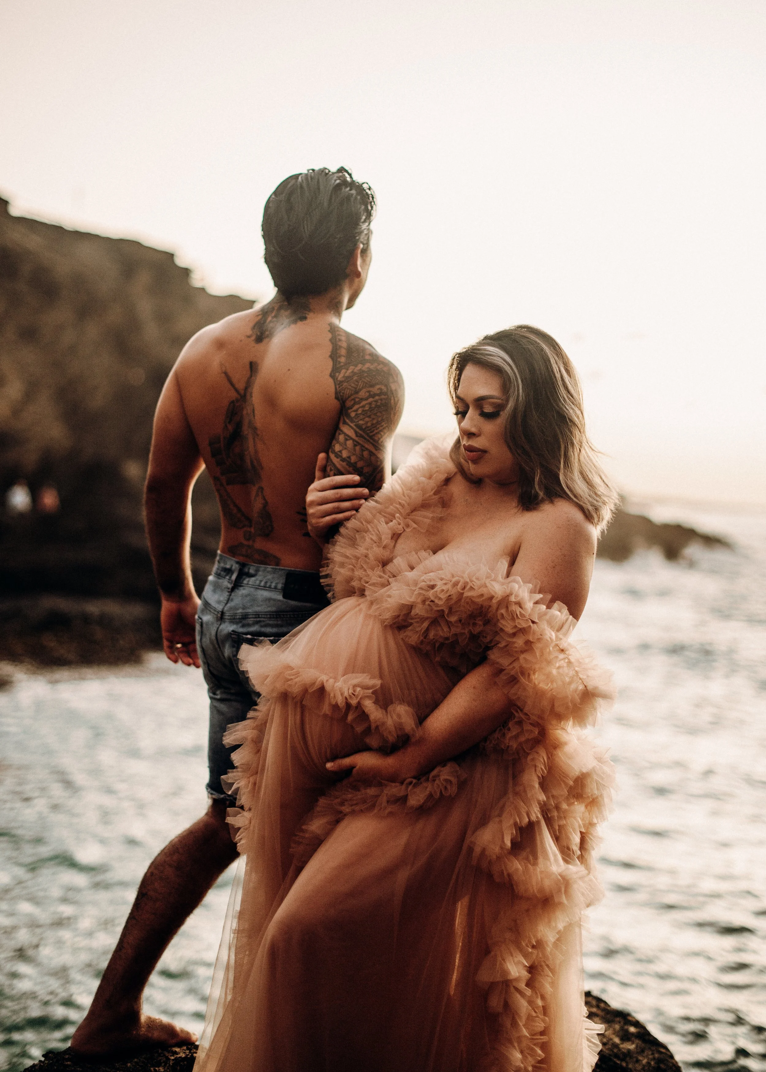 A man with tattoos on his back and arms and a woman in a pink, ruffled dress standing near the water against a rocky background at sunset.