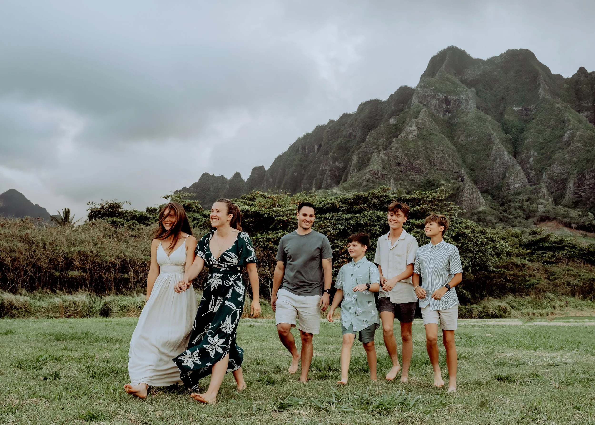 A group of eight people walking outdoors on a grassy field with lush greenery and mountains in the background. The group consists of young adults and children, all smiling and enjoying the walk.