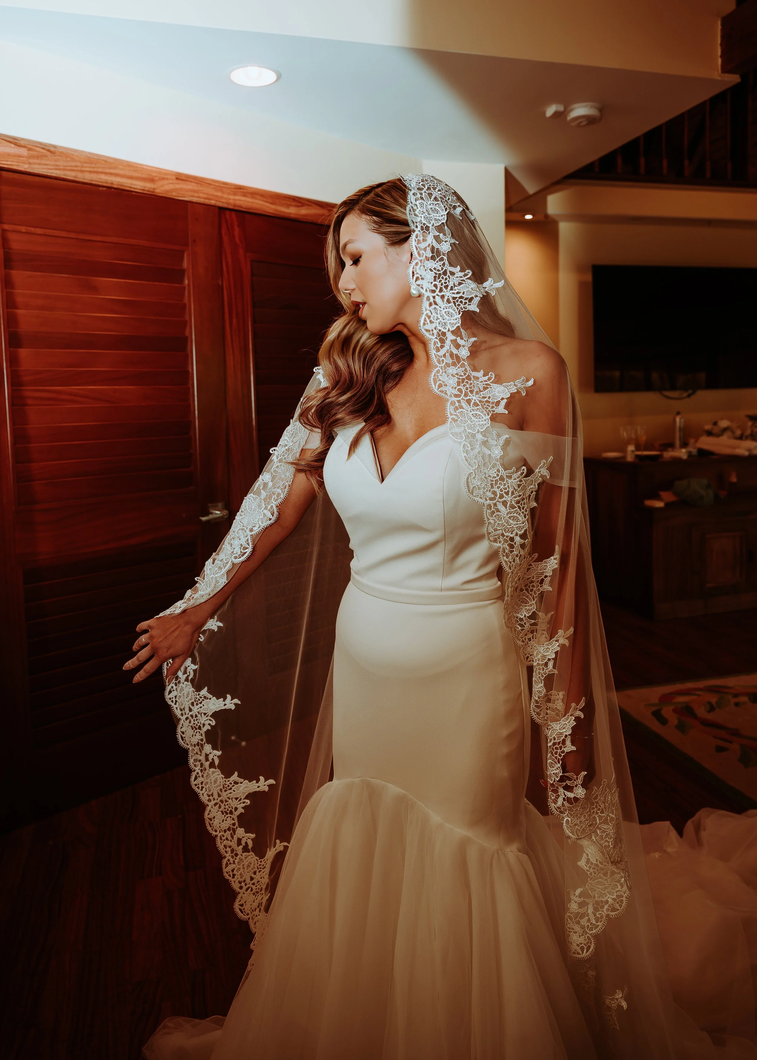 A bride in a white wedding dress with lace detailing on her veil, standing indoors with wooden paneling and a television in the background. | Best Hawaii Wedding Photographer & Videographer | Oahu, Hawaii Island, Kauai, and Maui 