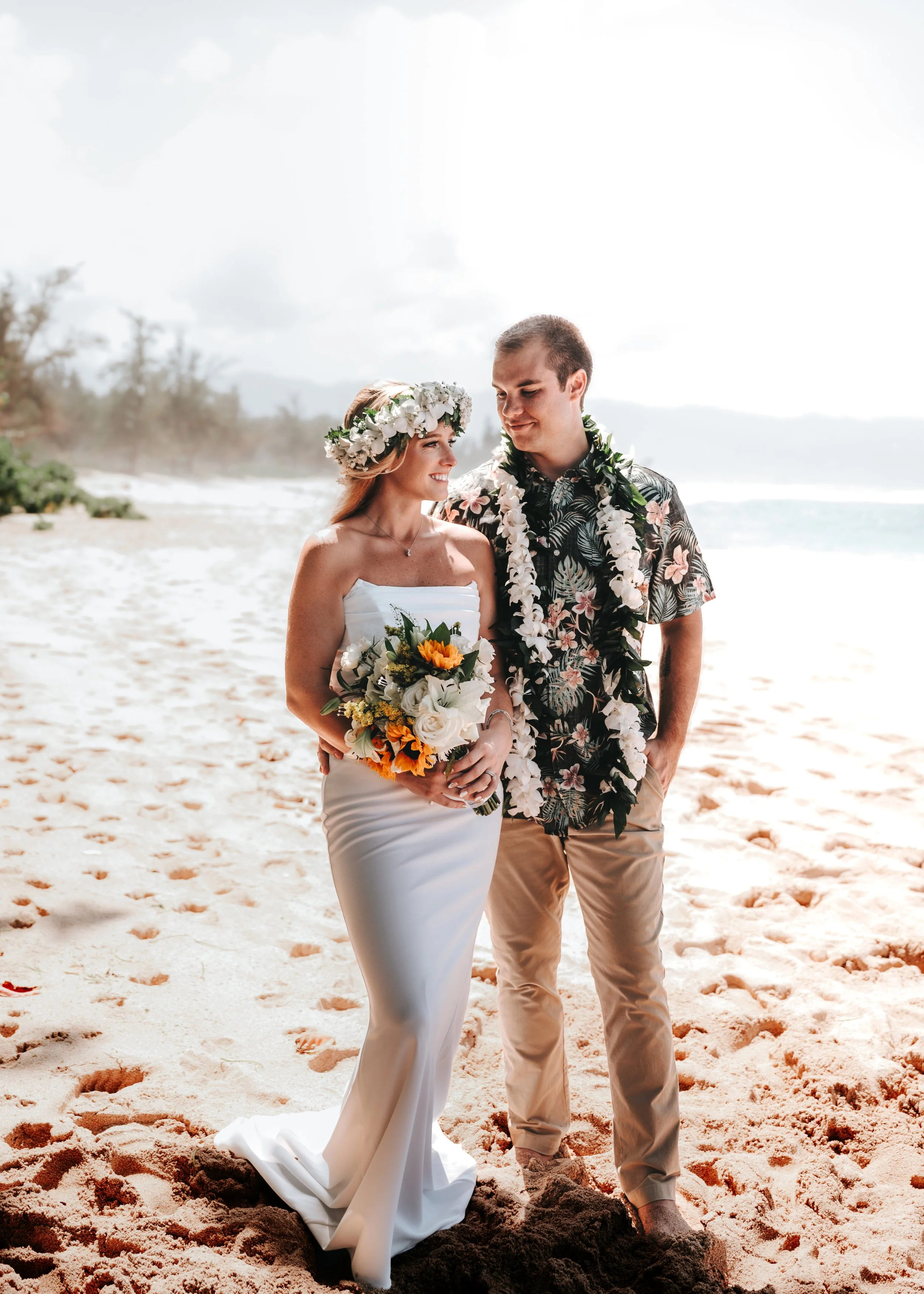 A bride and groom standing on the beach, facing each other and smiling, with the ocean and sky in the background. The bride is wearing a white strapless wedding gown and a floral crown, holding a bouquet of flowers. The groom is wearing a tropical print shirt, white lei, and beige pants.