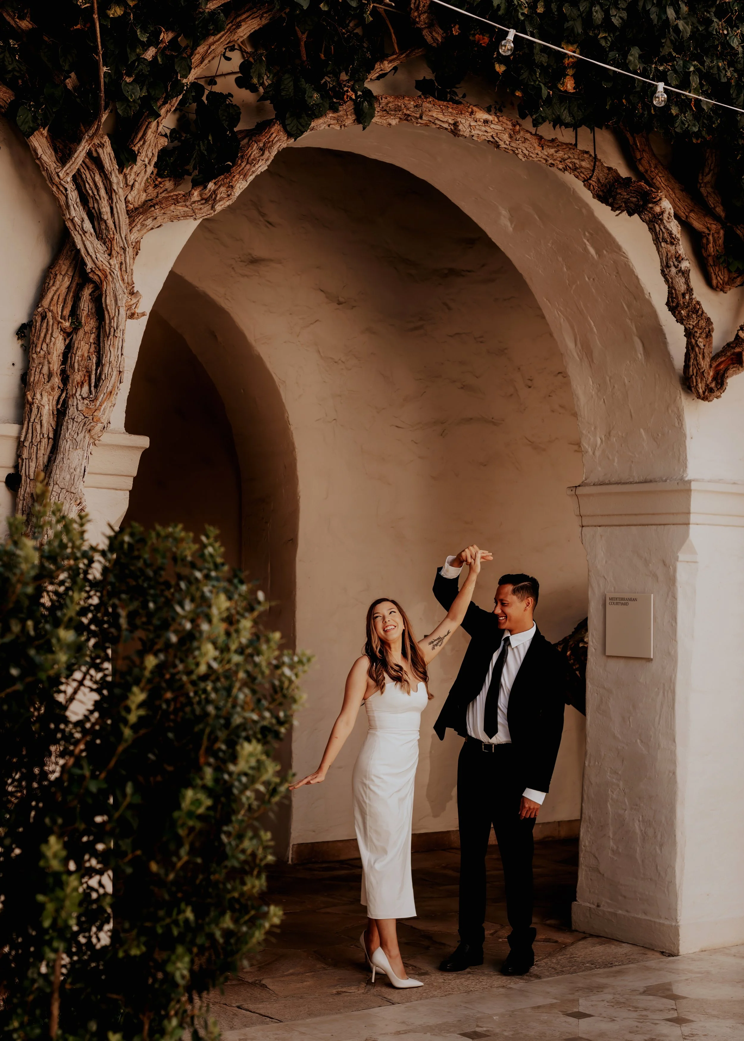 A bride in a white dress and high heels dancing with a groom in a black suit, white shirt, and black tie in an arched outdoor corridor decorated with greenery and string lights.  | Best Hawaii Wedding Photographer & Videographer | Oahu, Hawaii Island