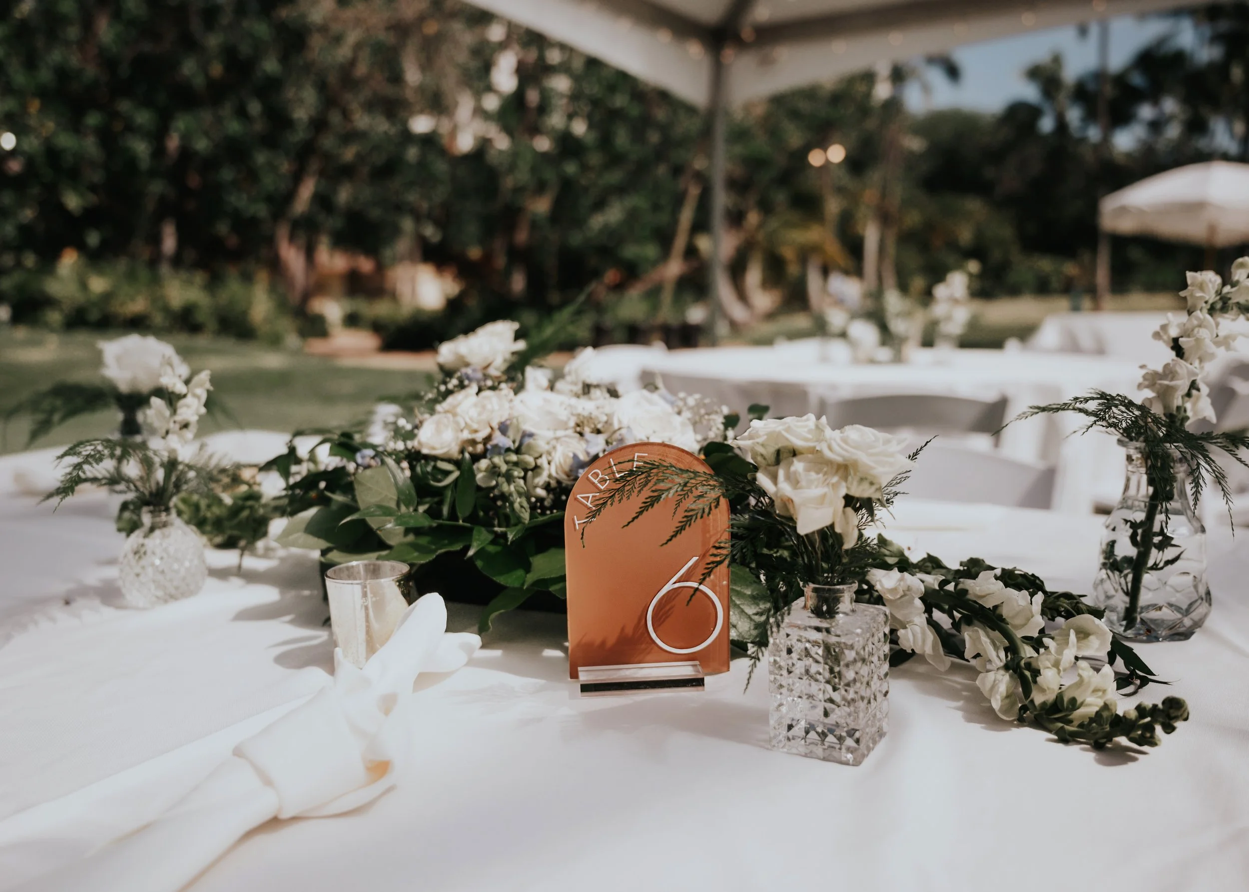 Wedding reception table decorated with white flowers, glass vases, and table number 2, outdoors under tents with trees in the background. | Best Hawaii Wedding Photographer & Videographer | Oahu, Hawaii Island, Kauai, and Maui 