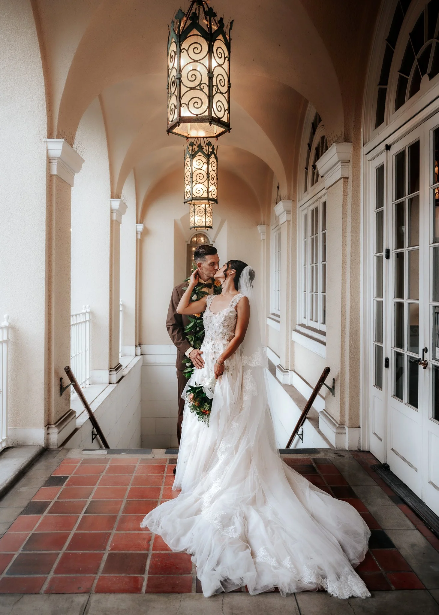 A bride and groom sharing a kiss in a covered outdoor corridor with hanging lanterns and arched windows. | Best Hawaii Wedding Photographer & Videographer | Oahu, Hawaii Island, Kauai, and Maui 