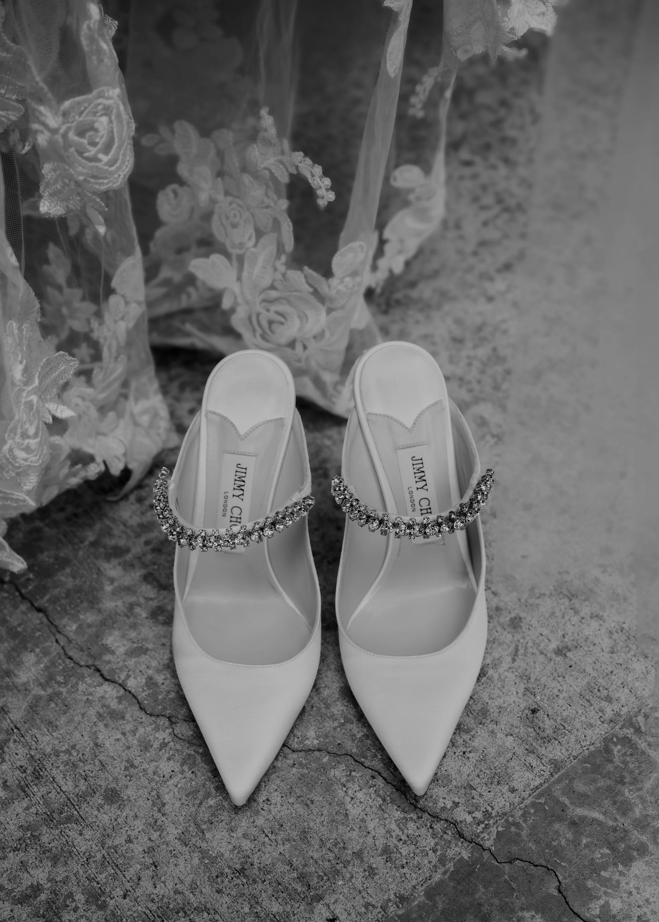 A pair of white pointed-toe high heels with rhinestone ankle straps, viewed from above, on a rough concrete surface. The shoes have the label 'Jimmy Choo London' inside.  | Best Hawaii Wedding Photographer & Videographer | Oahu, Hawaii Island, Kauai,