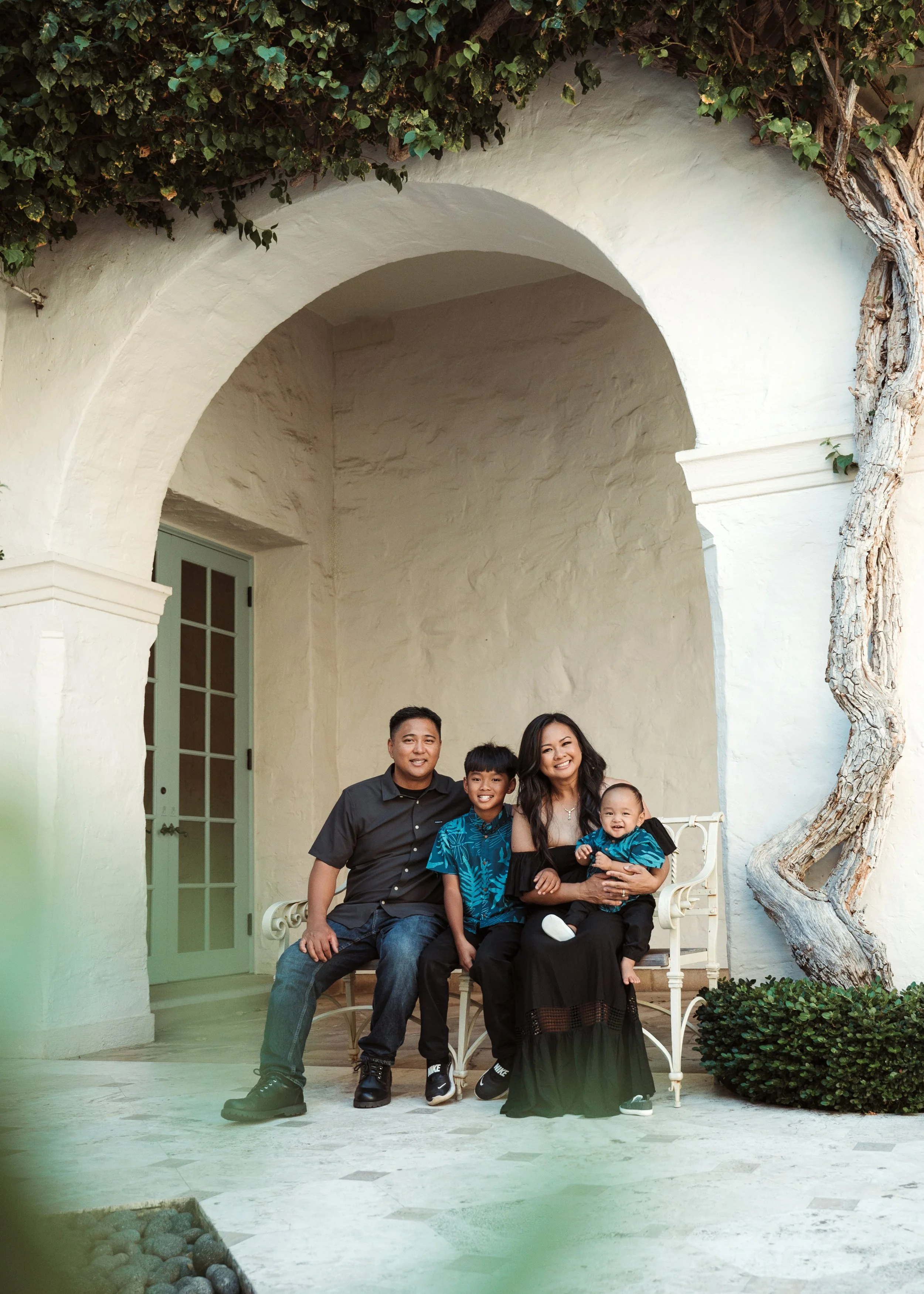 A family of five sits on a white bench under a white archway with green door and window behind them, surrounded by greenery including a large tree with twisted branches.