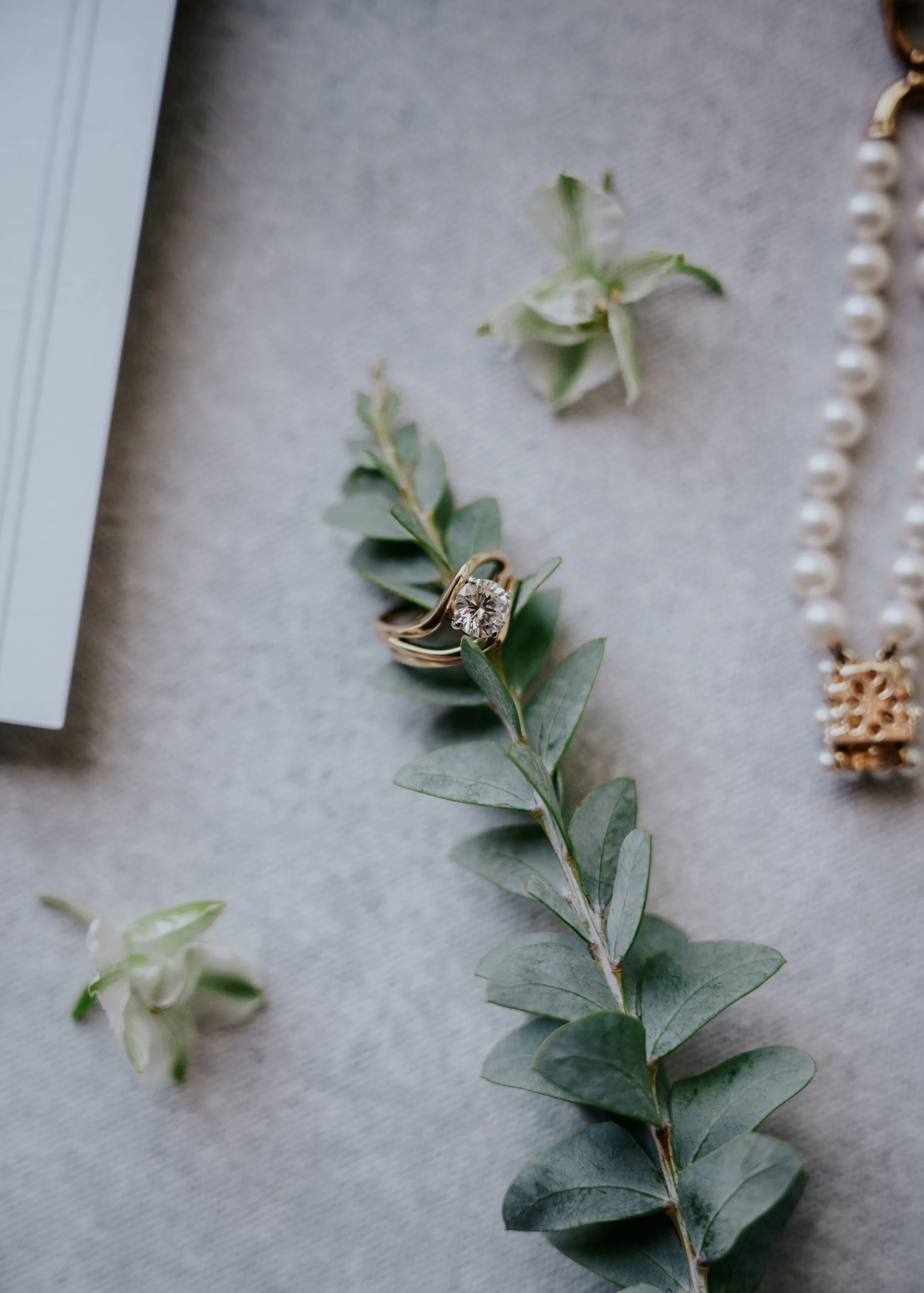 A branch of eucalyptus leaves adorned with a gold ring featuring a large, faceted gemstone and a string of pearls, placed on a light-colored fabric surface. | Best Hawaii Wedding Photographer & Videographer | Oahu, Hawaii Island, Kauai, and Maui 